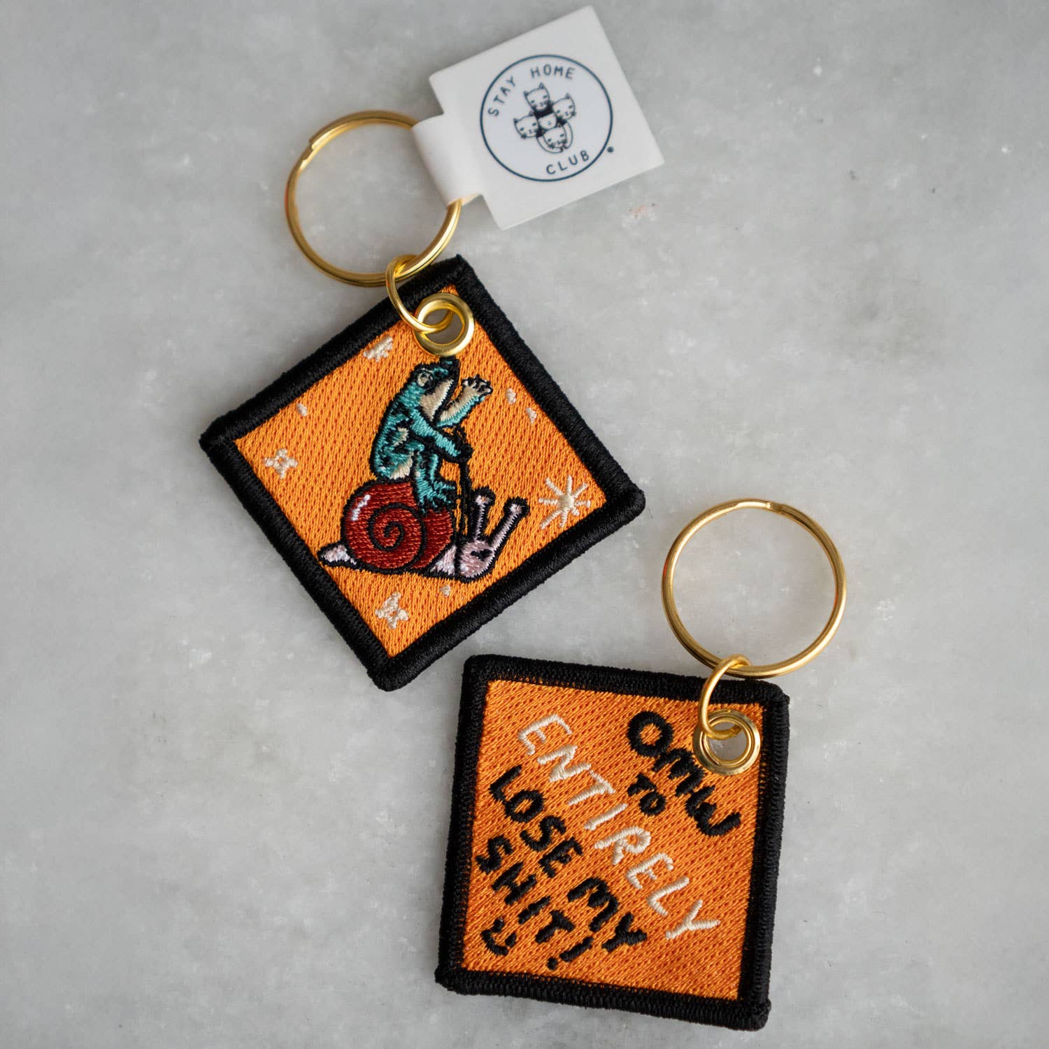 Two square keychains with embroidered designs on a gray surface, featuring a brand tag.