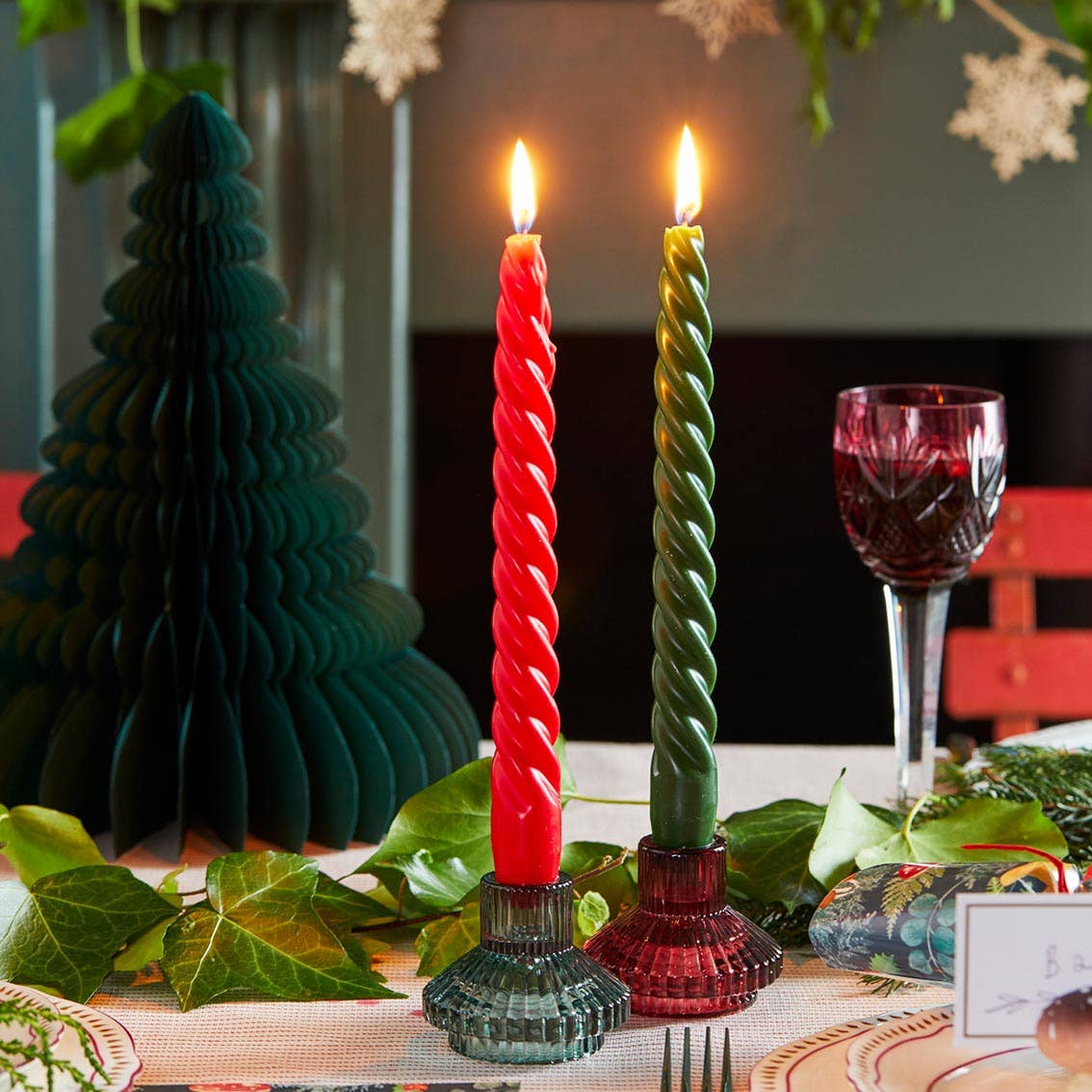 Twisted red and green candles on a festive table with a Christmas tree decoration.