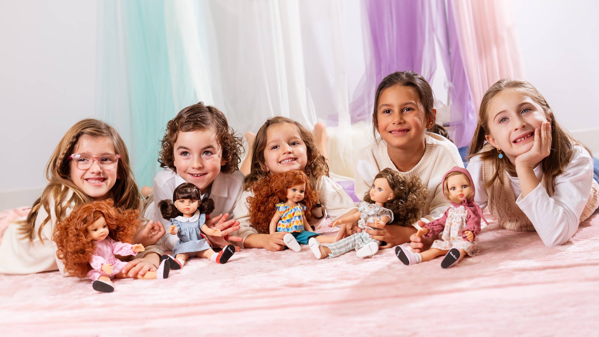 Six young girls lying side-by-side on a pink blanket, each holding a small doll. They are posed in front of a soft, draped pastel background of white, mint green, and lavender.