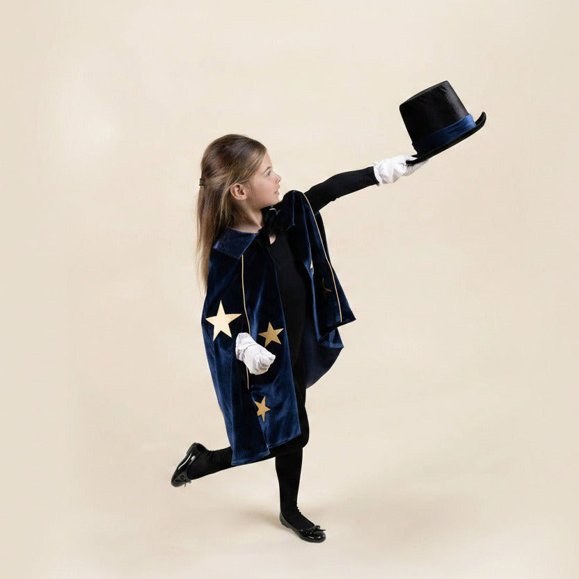A young girl in a magician costume poses dynamically against a solid beige background, wearing a navy blue velvet cape with gold stars, a black dress, white gloves, and holding a black top hat.