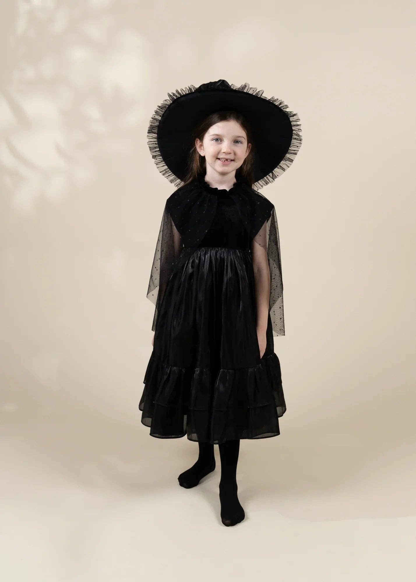 A young girl modeling a black dress costume with sheer cape sleeves, a wide-brimmed black hat with ruffled trim, and black tights, standing against a warm beige studio background.