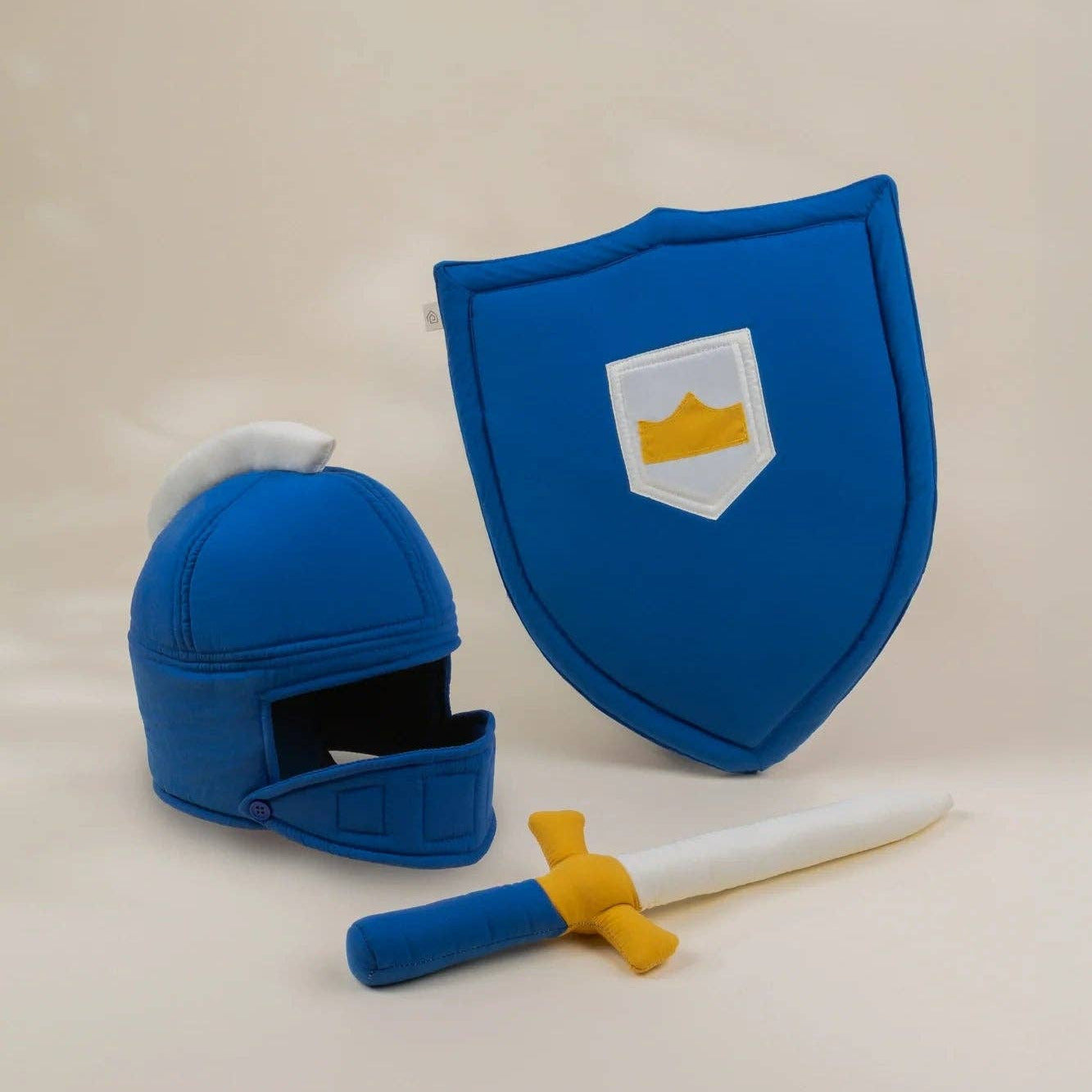 A miniature medieval knight toy set consisting of a blue helmet with a white plume, a matching blue shield with a white and gold crest, and a small sword with a blue handle and gold crossguard, displayed on a light beige background.