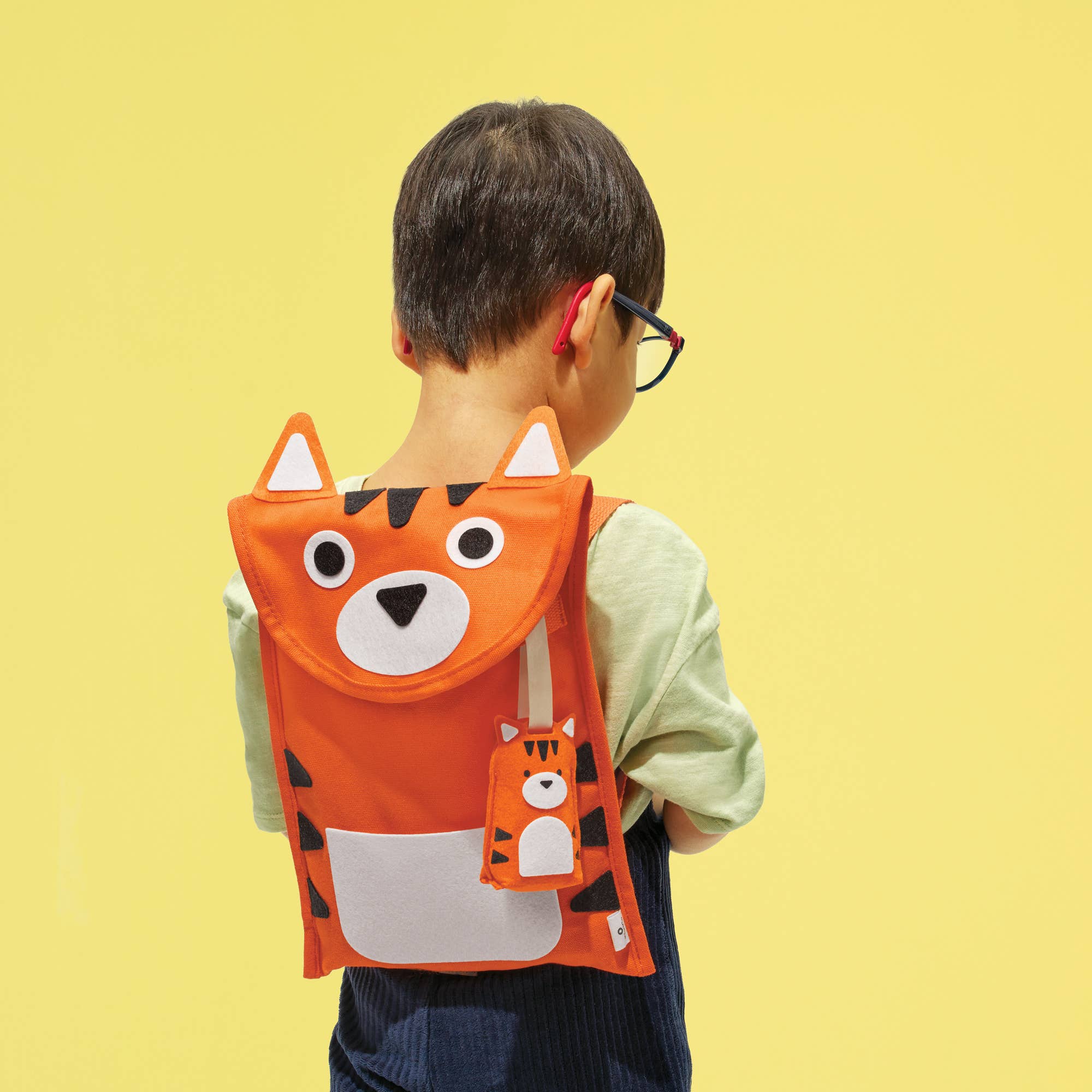 A child viewed from behind wearing a bright orange tiger-face backpack with black stripes, white inner ears, and a small matching tiger charm, against a solid pale yellow background.