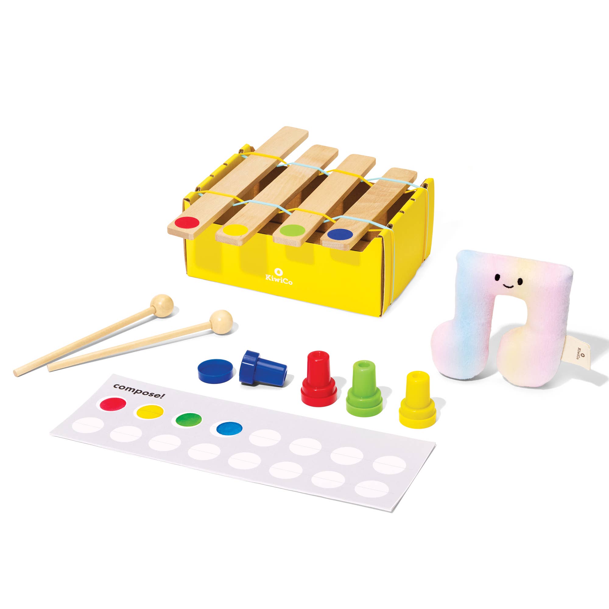 A children's wooden xylophone toy set with a yellow box, wooden mallets, colored percussion keys, a white composition sheet, and a pastel iridescent U-shaped pillow on a white background.