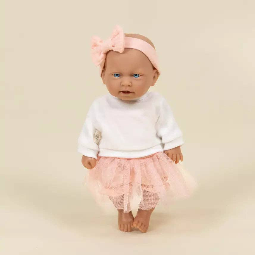 A baby doll with realistic skin and blue eyes, wearing a white long-sleeve top, a light pink ruffled skirt, and a matching pink bow headband, standing against a plain beige background.