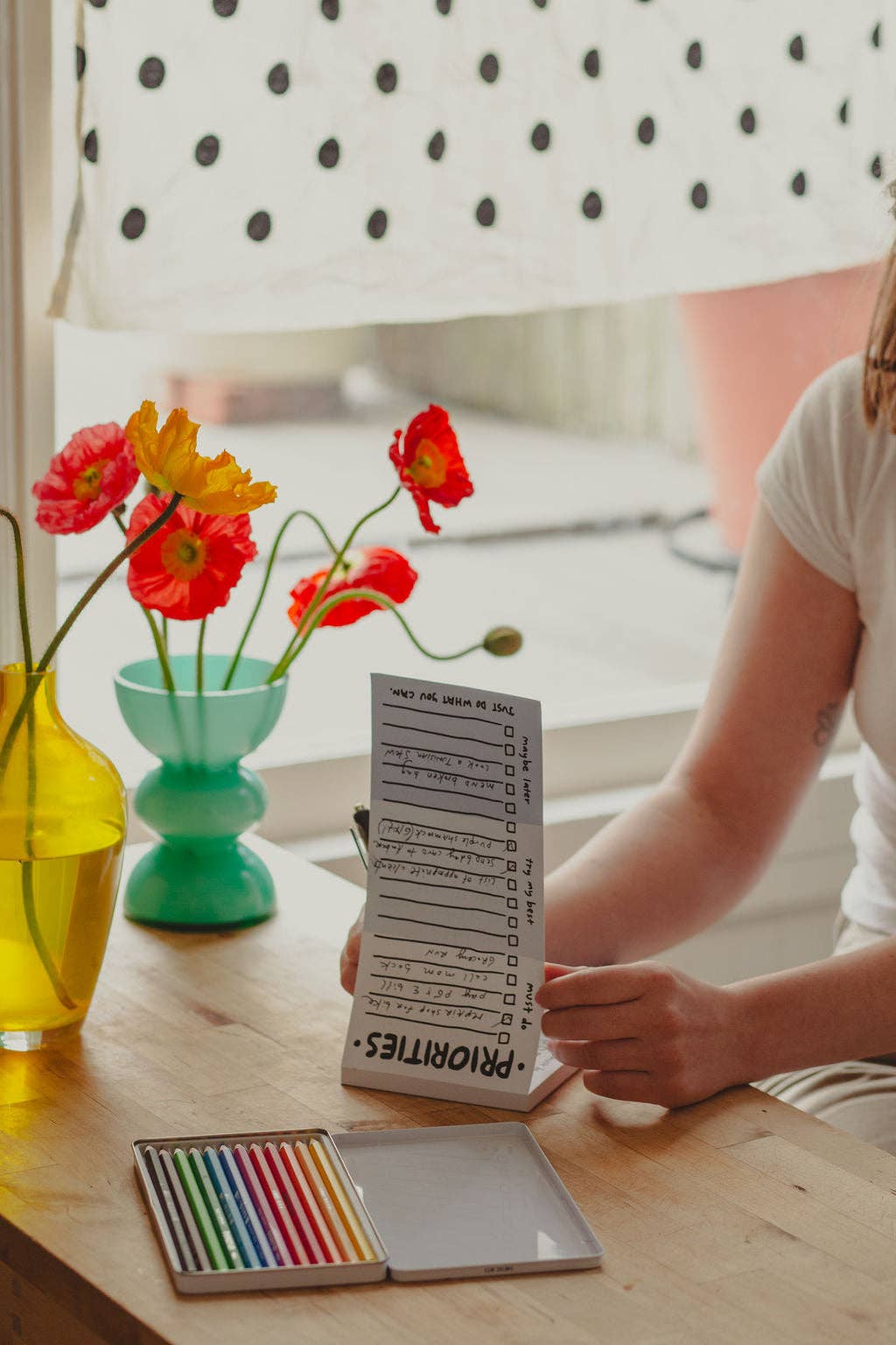 Person holding a notepad with a list, surrounded by flowers and art supplies on a table.