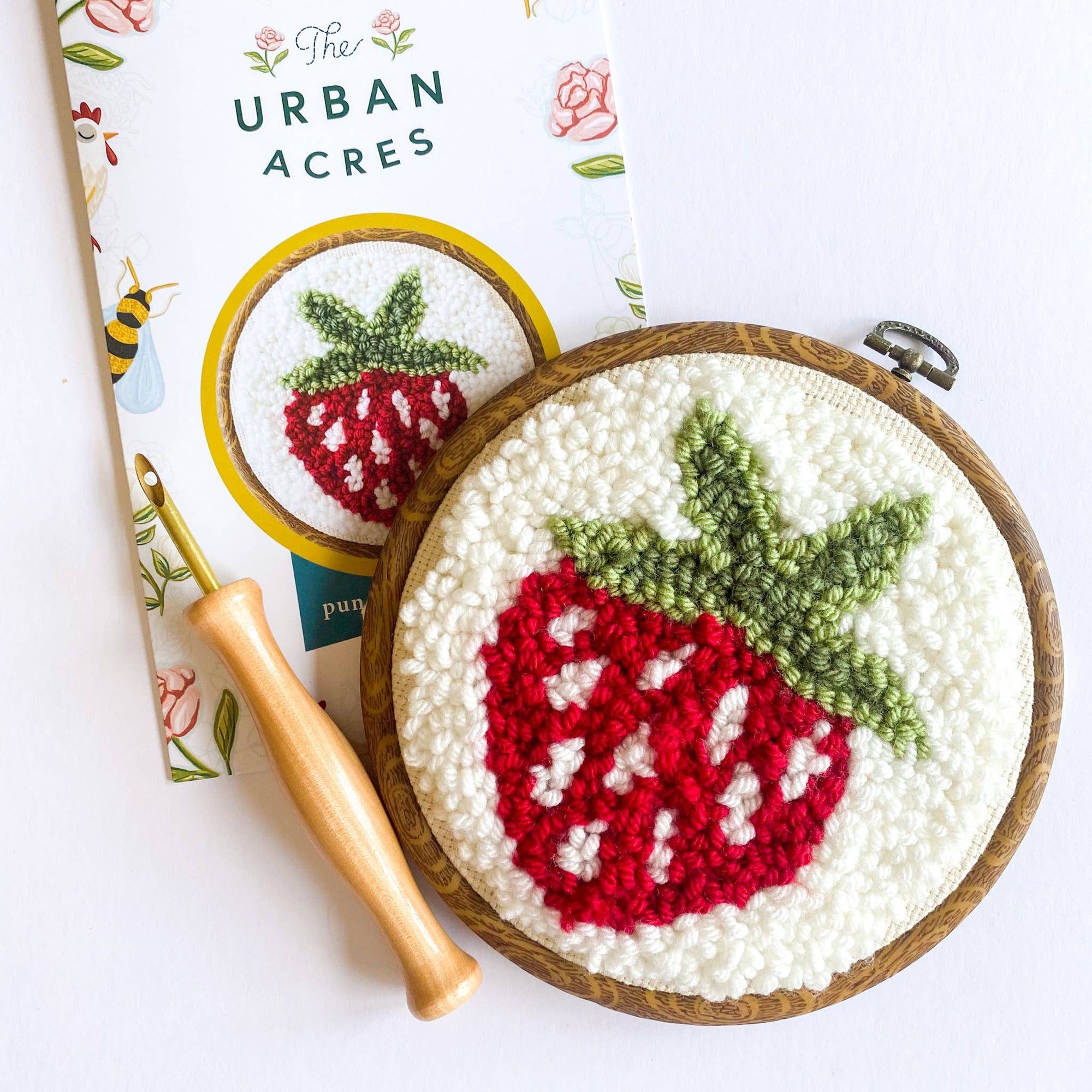 Embroidery hoop with strawberry design next to a book titled 'The Urban Acres' on a white background