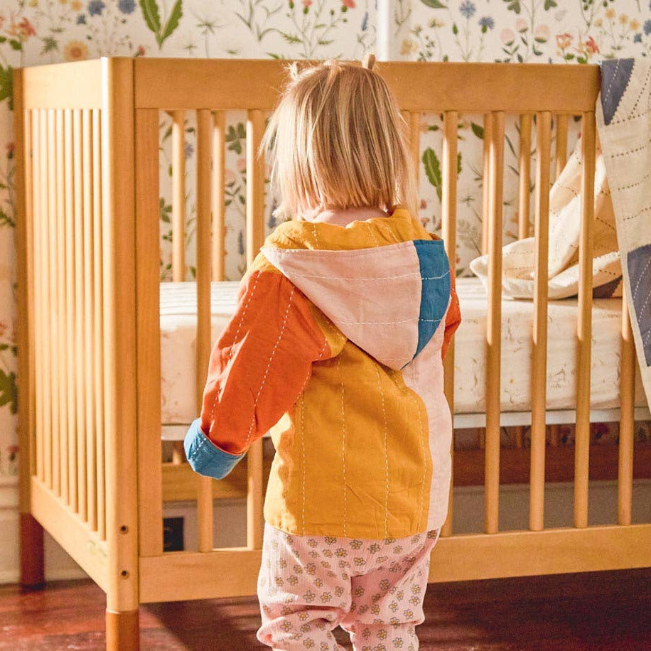 Child standing in a nursery with a wooden crib and colorful rug.