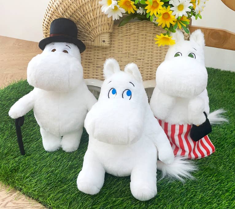 Three white plush toys with different accessories on a grass-like surface with flowers in the background.