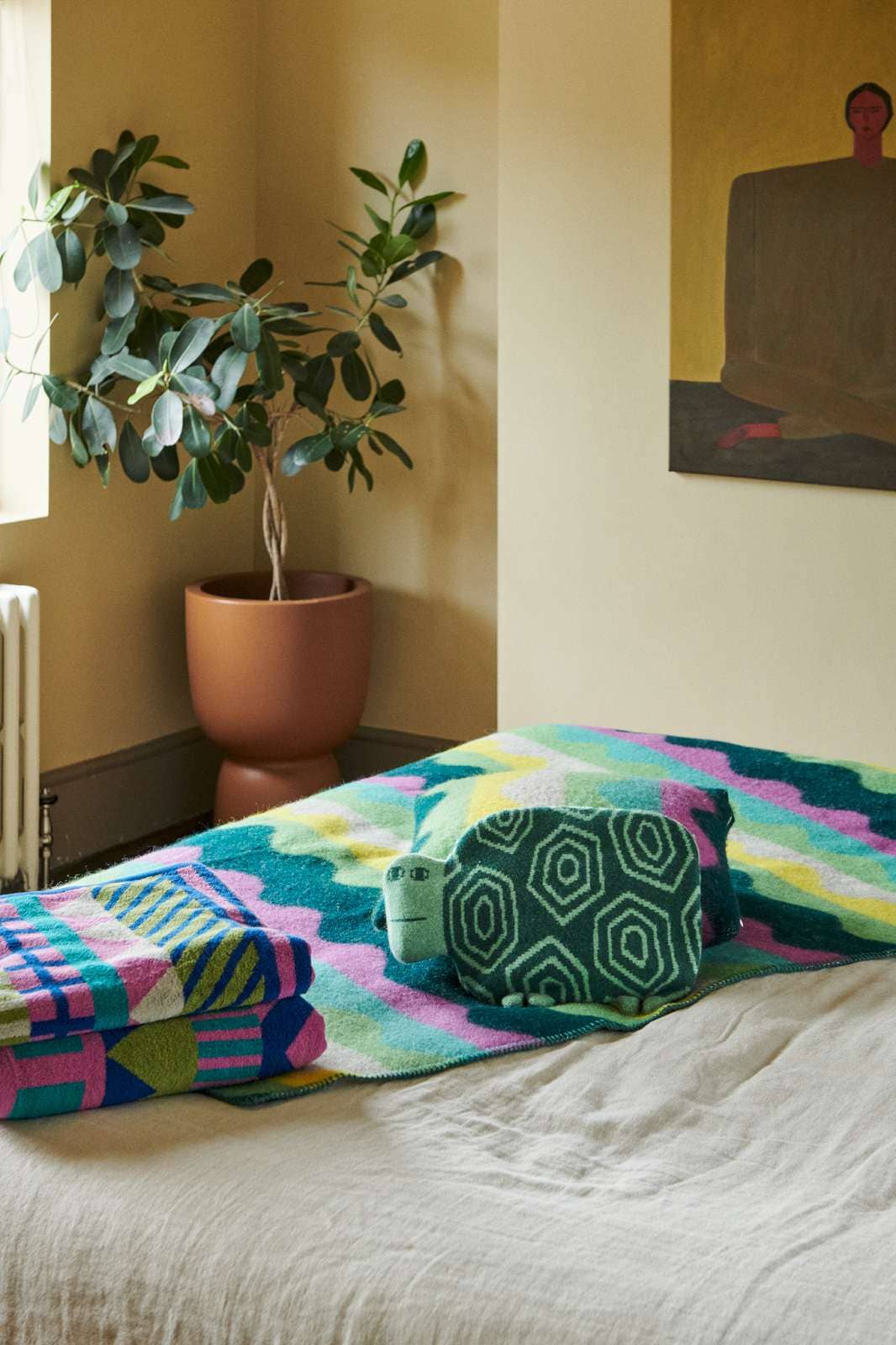 A colorful patterned throw blanket and matching pillow set styled on a bed in a warm, sunlit room with a potted plant and wall art in the background.