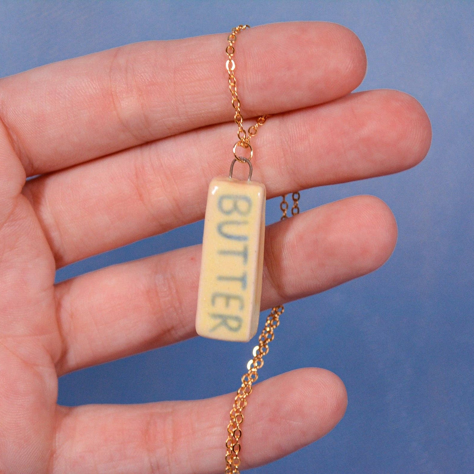 Hand holding a necklace with a 'BUTTER' pendant against a blue background