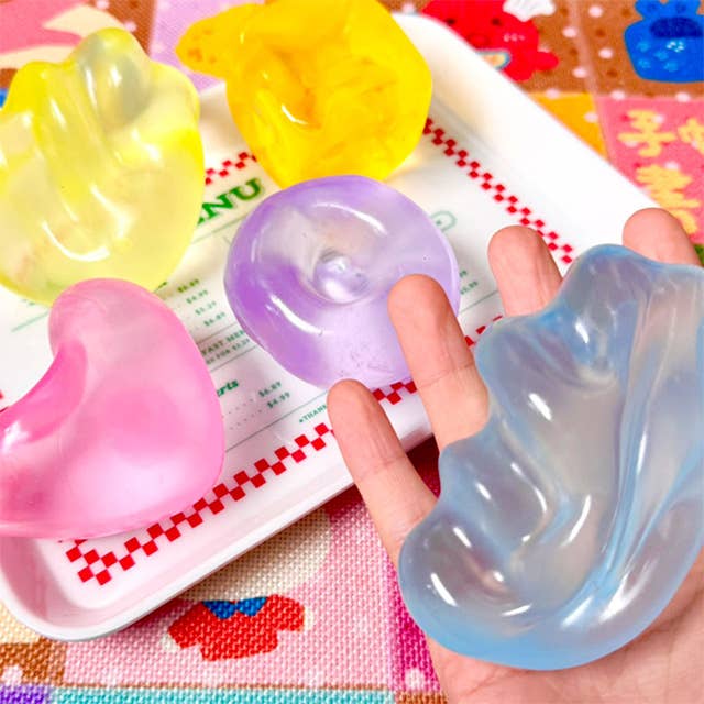 A hand holds a translucent light blue gel toy, with other translucent gel ear toys in pink, purple, and yellow resting on a white tray in the background.