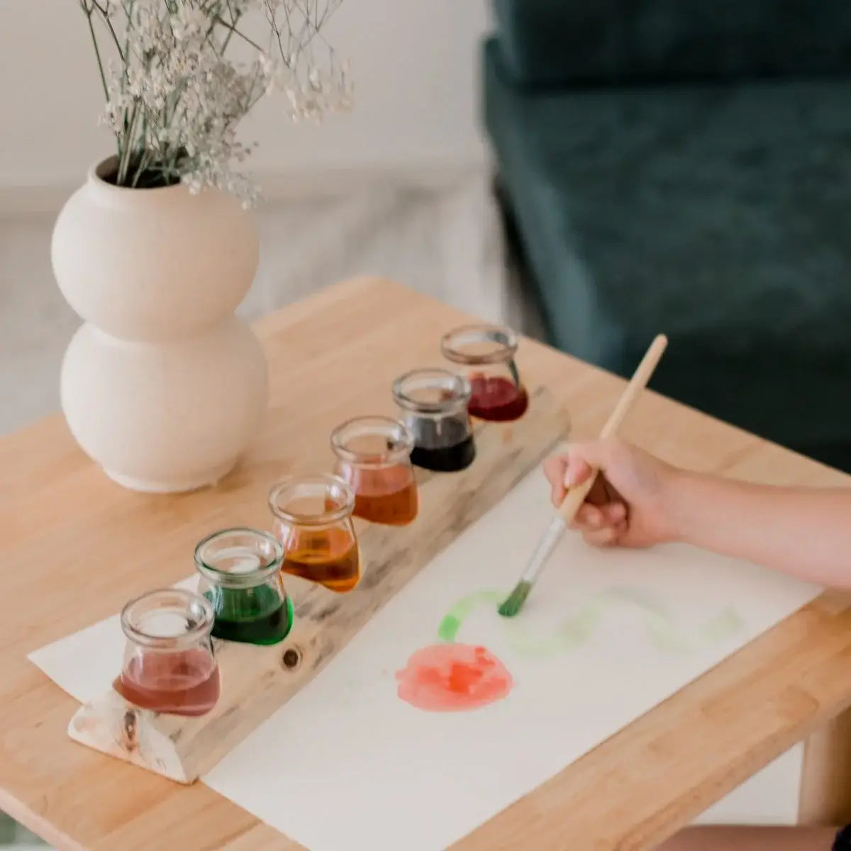 Person painting a flower on paper with watercolor paints on a wooden table.