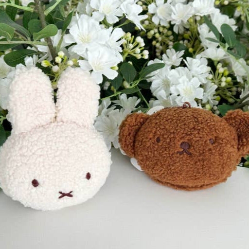Two small plush toys, one white rabbit and one brown bear, surrounded by white flowers and green leaves on a light surface.