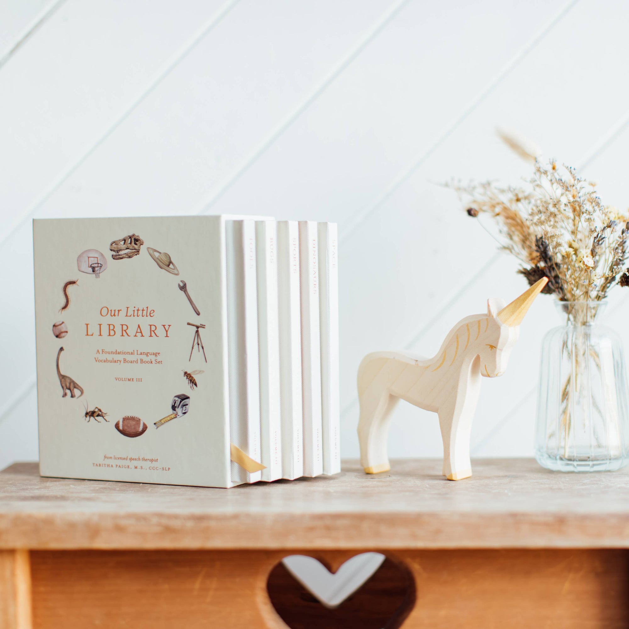Book titled 'Our Little Library' on a wooden shelf with a unicorn figurine and vase of dried flowers.