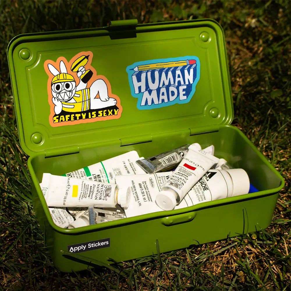A green metal storage case sitting outdoors on grass, open to reveal paint tubes inside. The inside lid has two stickers: one cartoon rabbit wearing a safety helmet with the text 'SAFETY IS SEXY' and another blue sticker that says 'HUMAN MADE'.