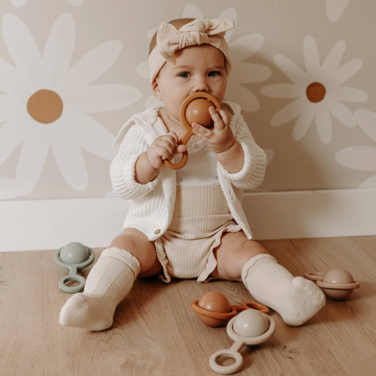 Baby sitting on a wooden floor with toys, wearing a white outfit and headband, against a floral wallpaper background.