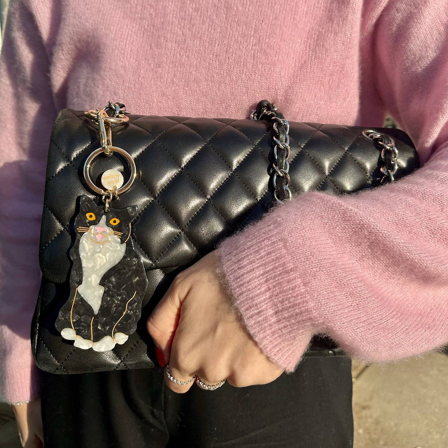 Person holding a black quilted handbag with a cat keychain, wearing a pink sweater.