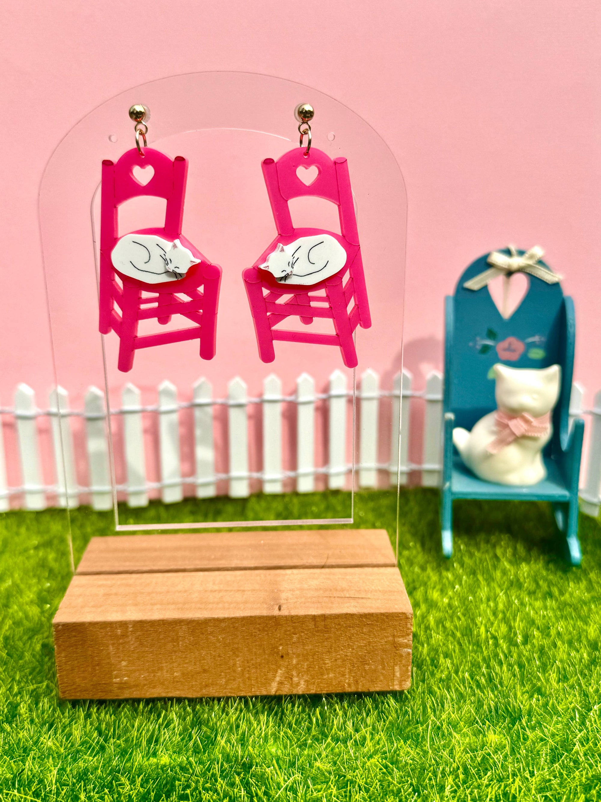 Pink chairs with white hearts on a clear stand against a pink background with a white picket fence and green grass.