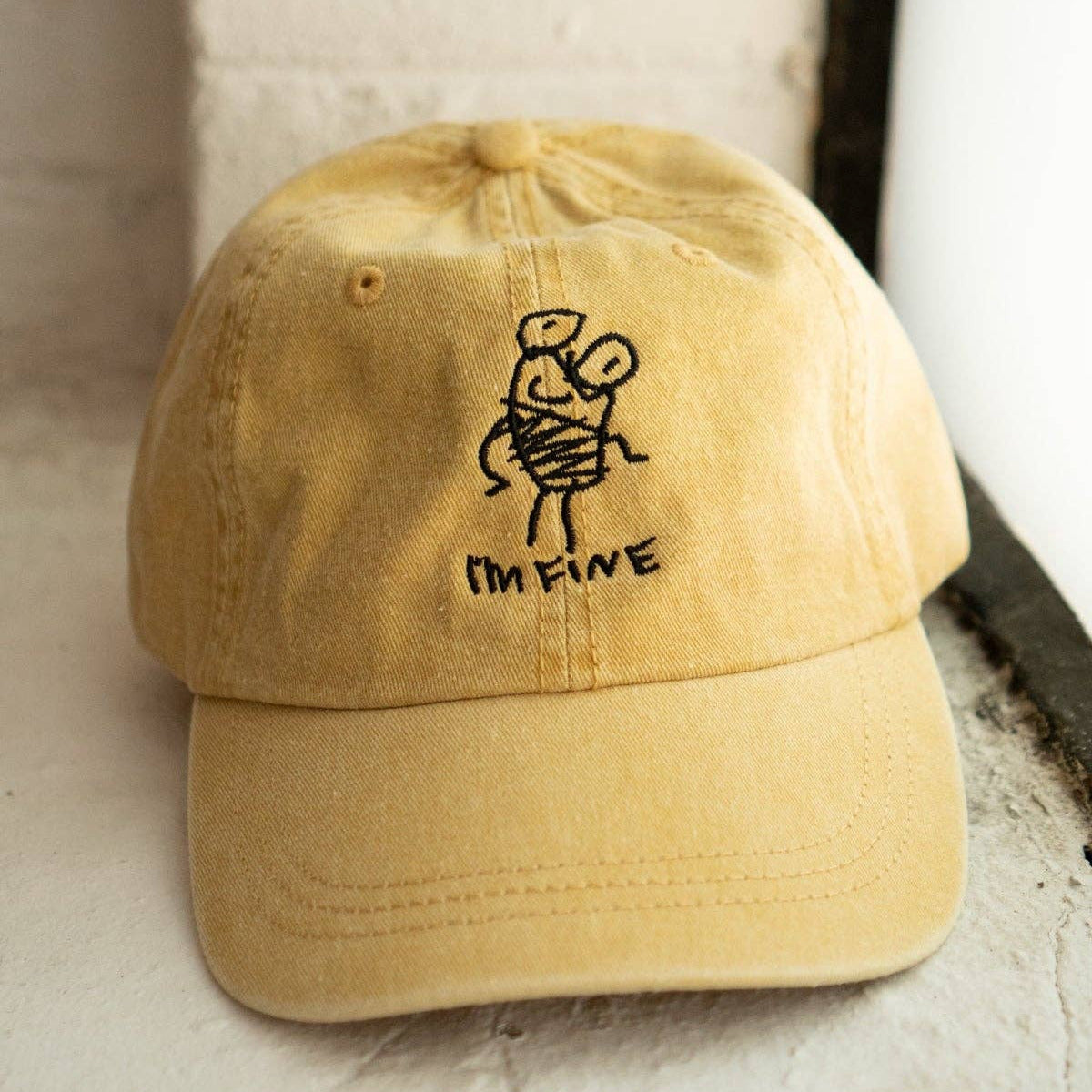 A yellow dad hat baseball cap with a black embroidered stick figure and the text 'I'M FIVE' on the front, resting on a textured white surface.