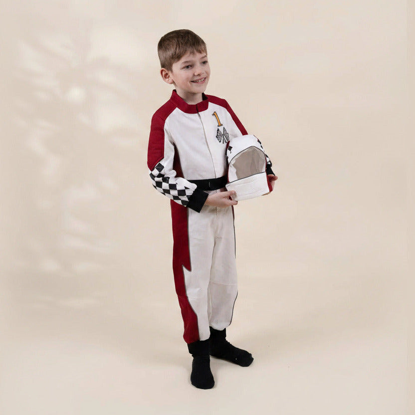 A young boy modeling a red and white racing suit costume, holding a white and black checkered racing helmet, against a solid beige studio background.