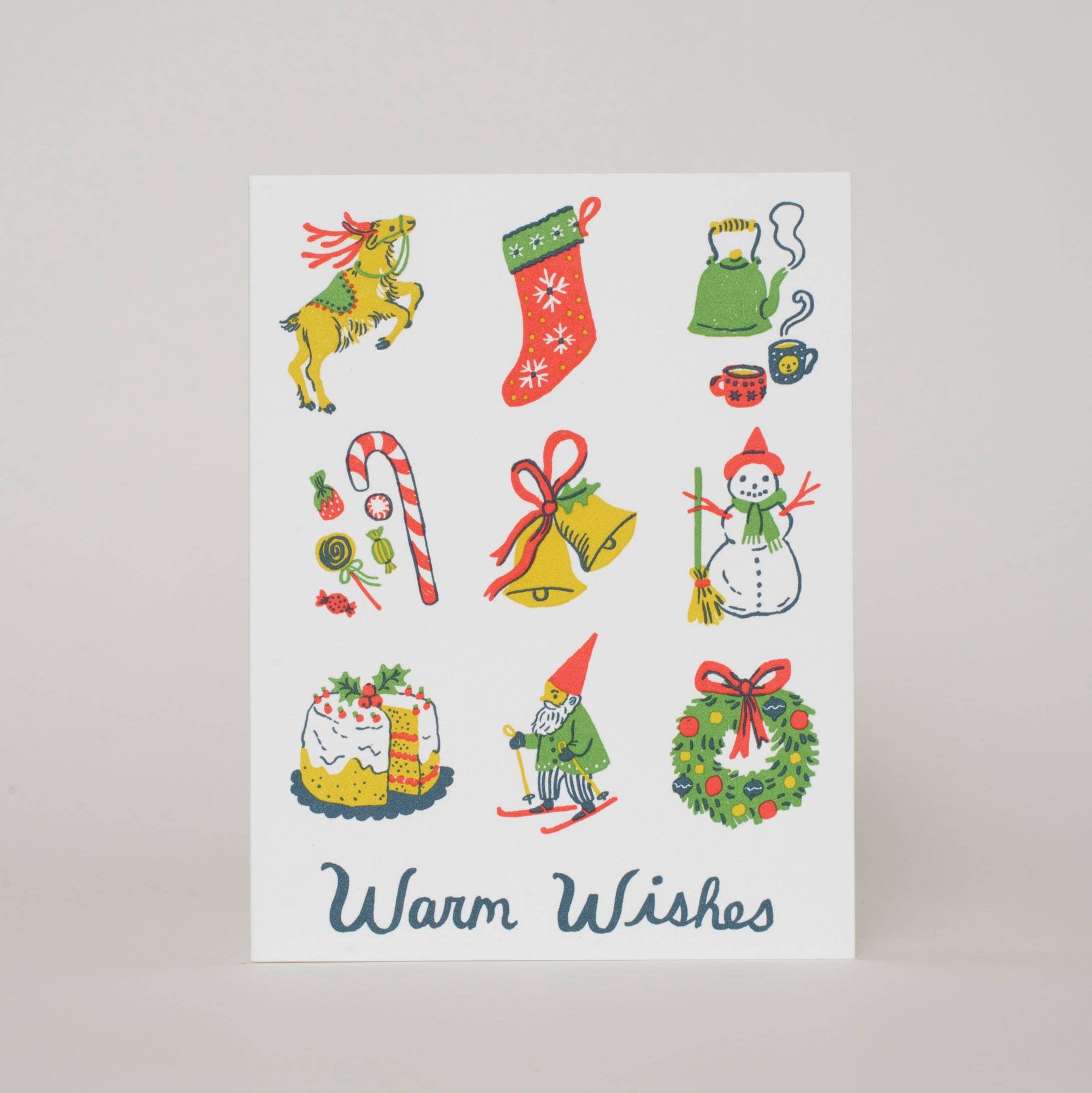 A vertical white greeting card featuring colorful Christmas-themed illustrations, including a reindeer, stocking, kettle, candy cane, bells, snowman, gnome, wreath, and cake, with the words 'Warm Wishes' in dark blue script at the bottom, photographed against a light gray background.