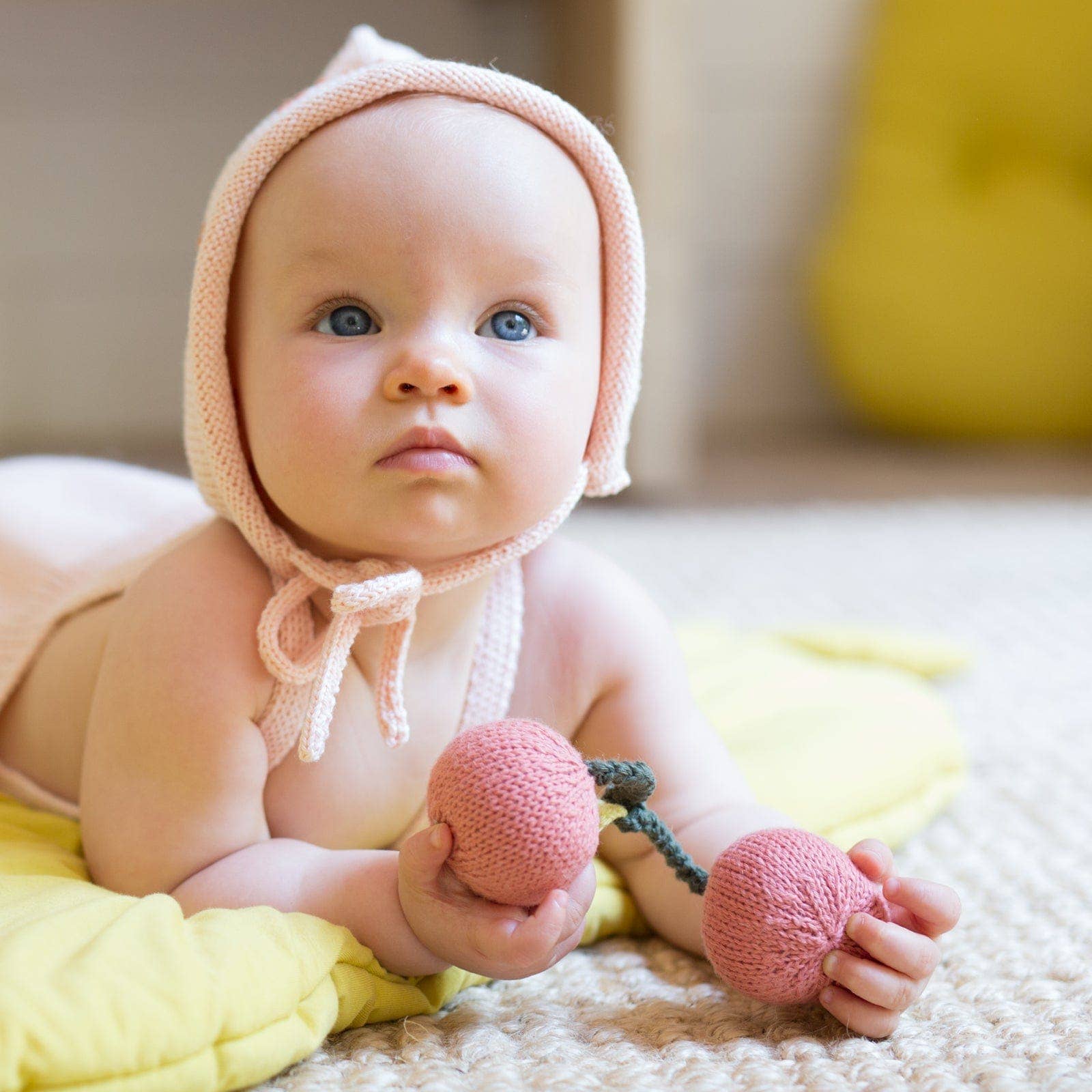 Baby wearing a pink knitted bonnet holding pink toys on a yellow blanket.