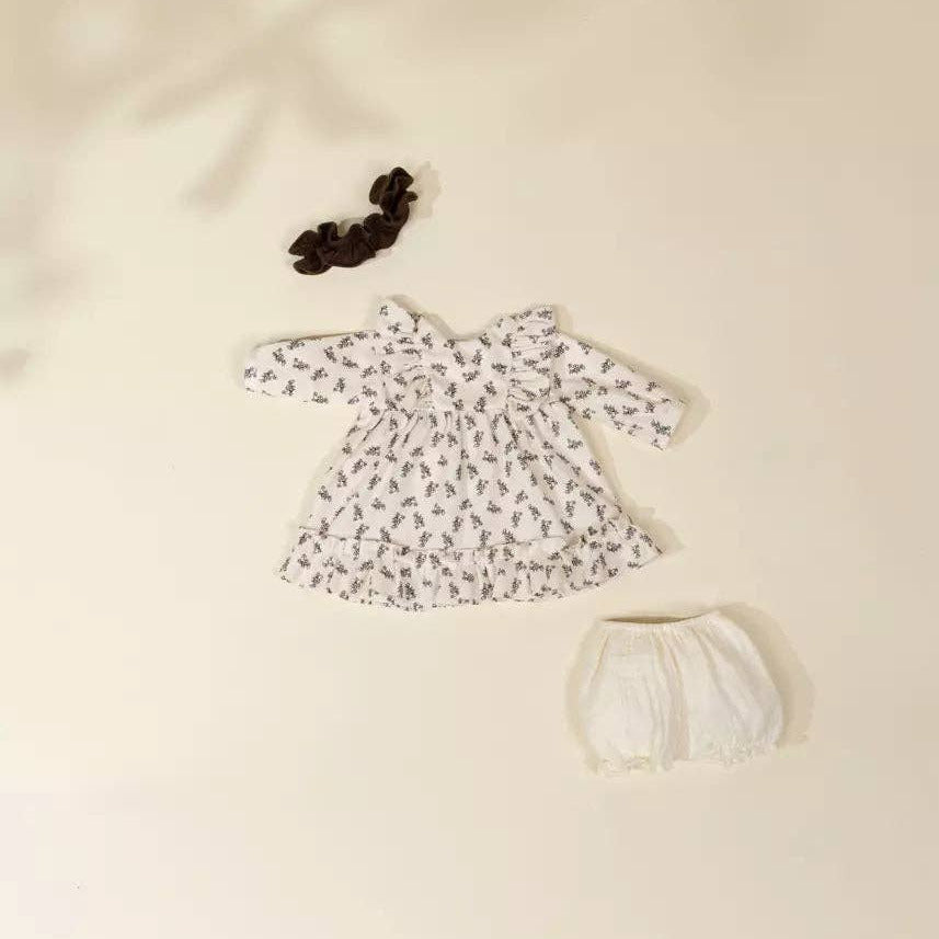 A baby outfit set laid flat on a warm cream background, including a long-sleeve white dress with a small dark floral print, white bloomers, and a dark scrunchie.