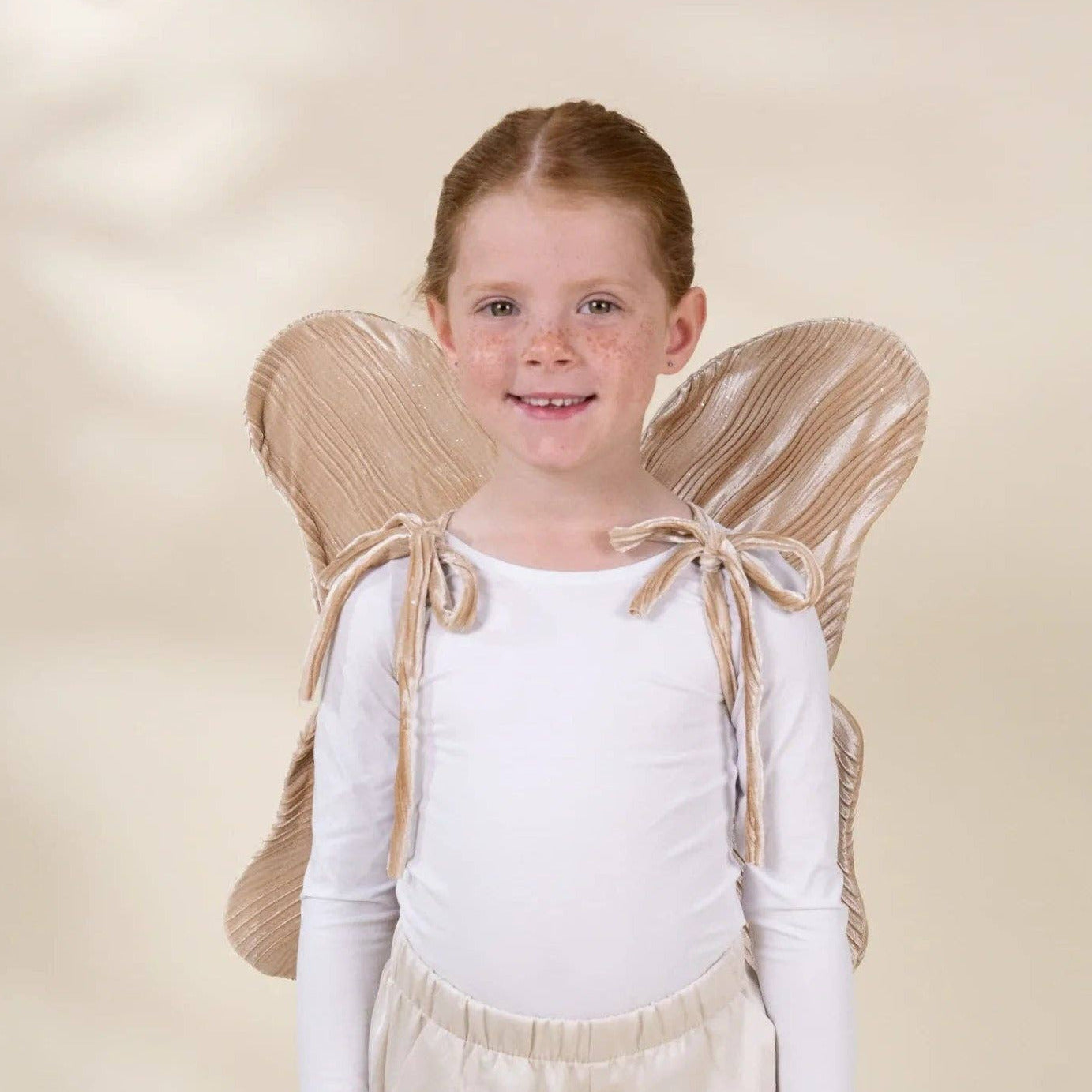 A young girl with light brown hair tied back stands against a warm beige studio background, wearing a white long-sleeve top, cream pants, and a beige angel costume with fabric wings and shoulder ties.