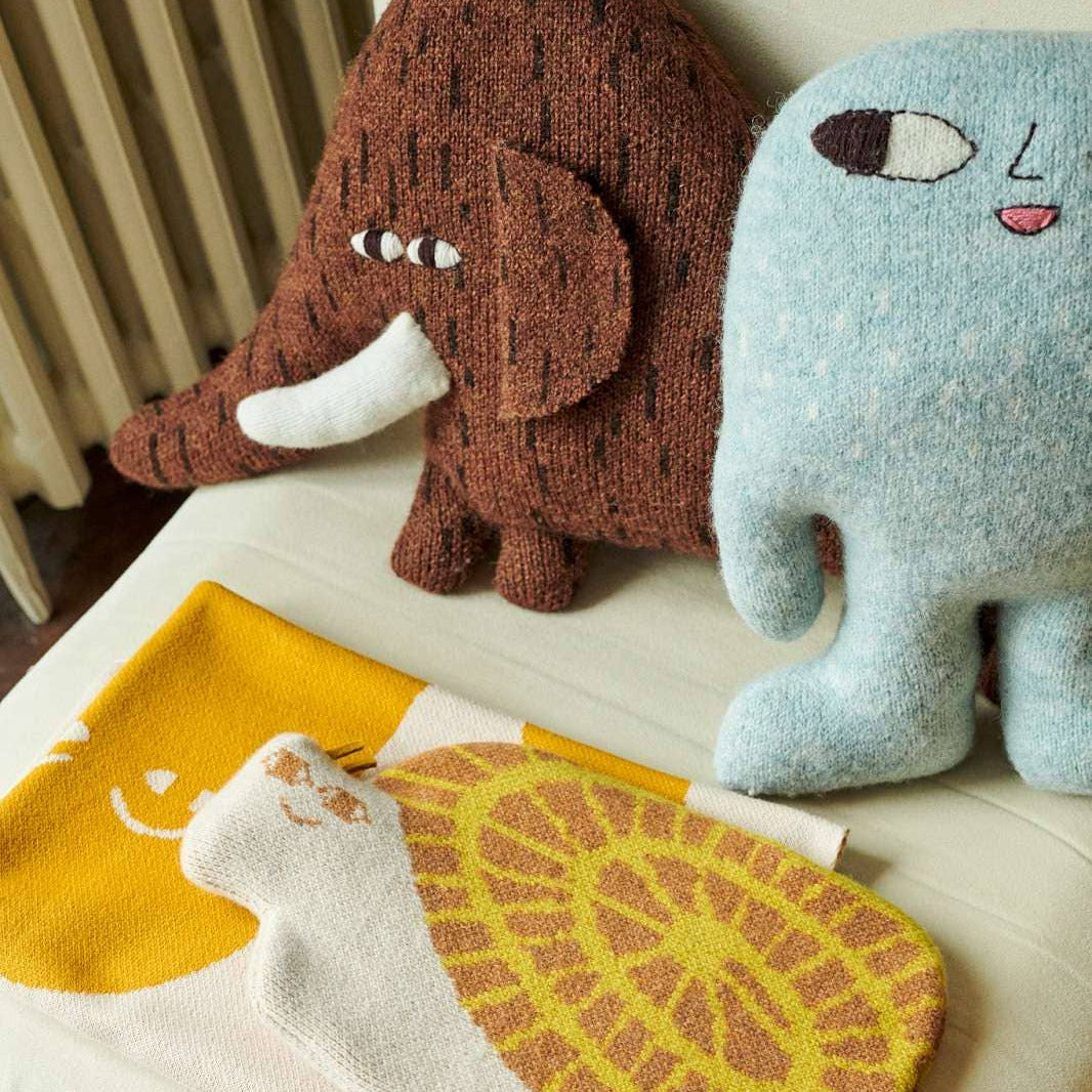 A cozy indoor scene showing a knitted plush elephant pillow in brown and a light blue plush elephant pillow on a cream cushion, with a yellow and cream patterned blanket or throw in front.