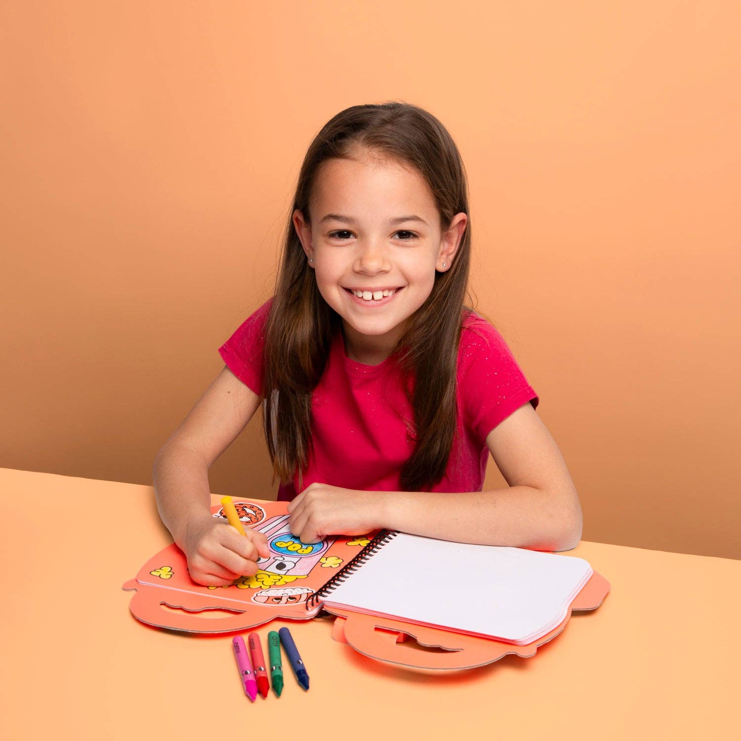 Young girl sitting at a table with coloring supplies against an orange background