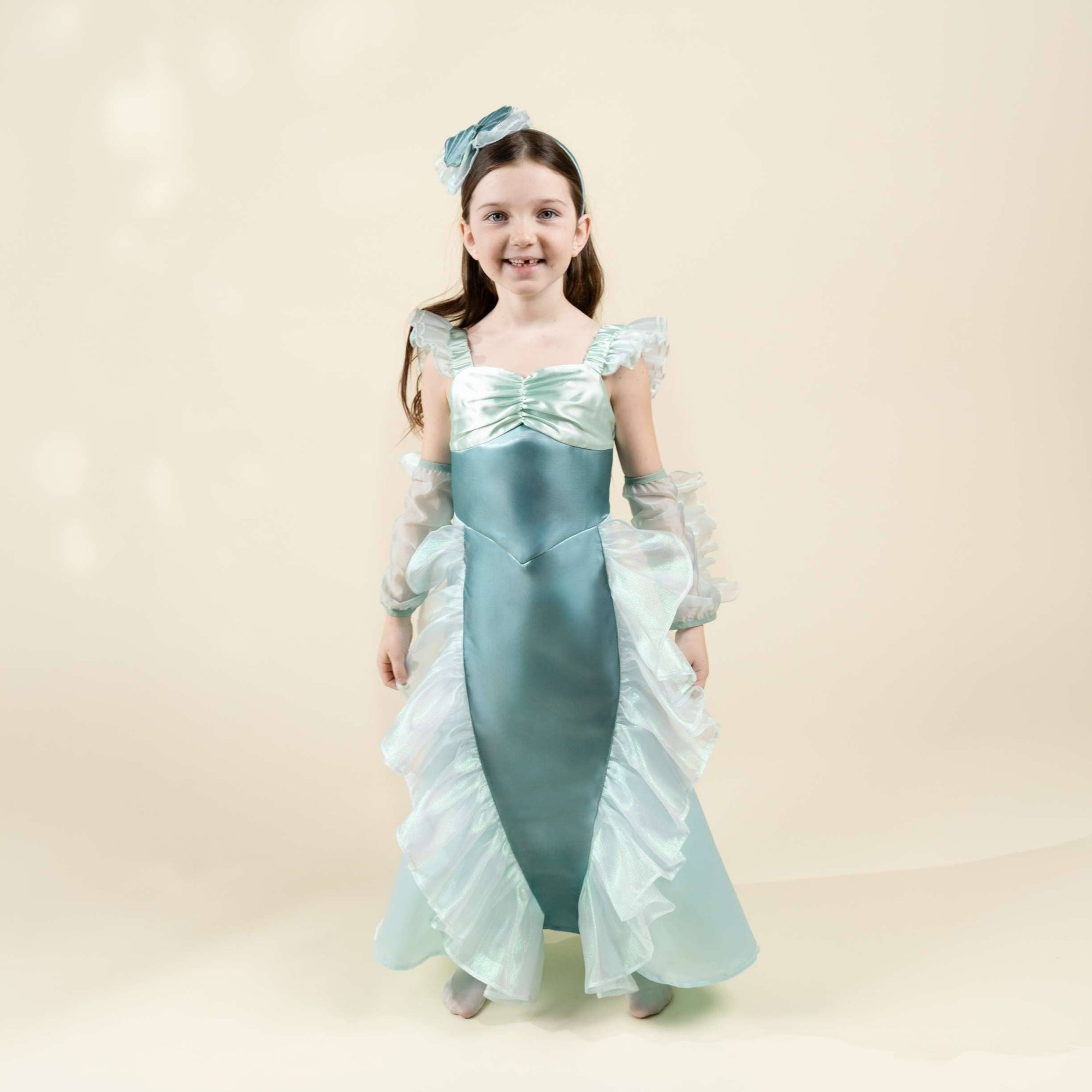 A young girl modeling a light blue and white princess-style costume dress with matching headpiece against a warm beige studio background.