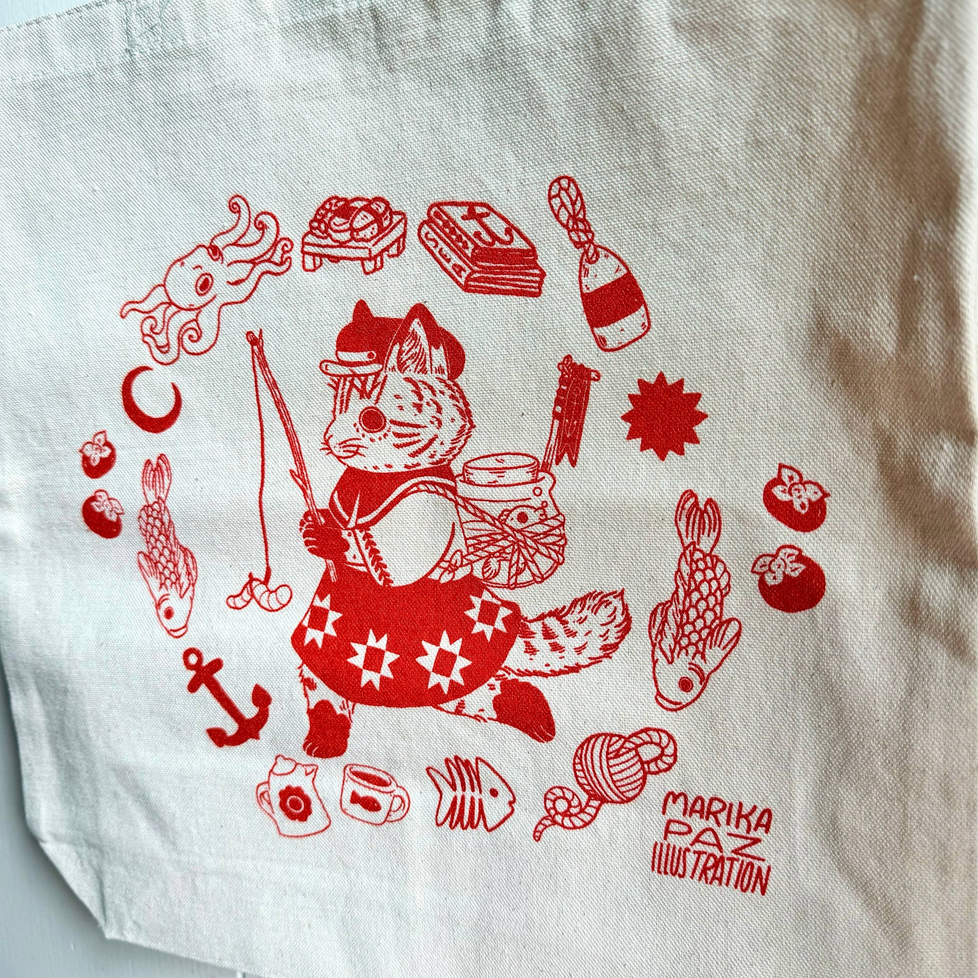 Beige tote bag with red illustrations and 'Marika Paz Illustration' text on a white background