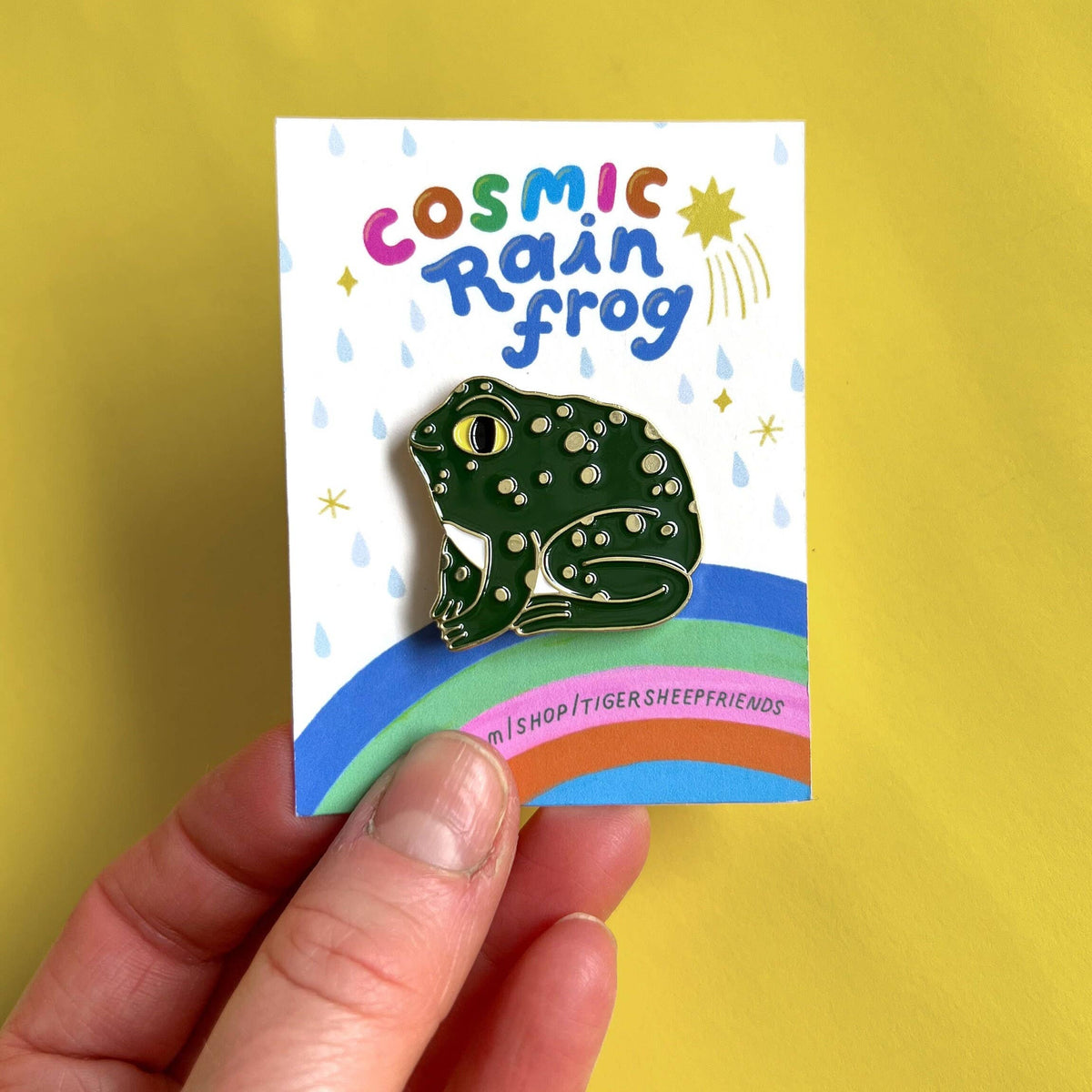 Pin with a frog design on a card labeled 'Cosmic Rain Frog' held against a yellow background