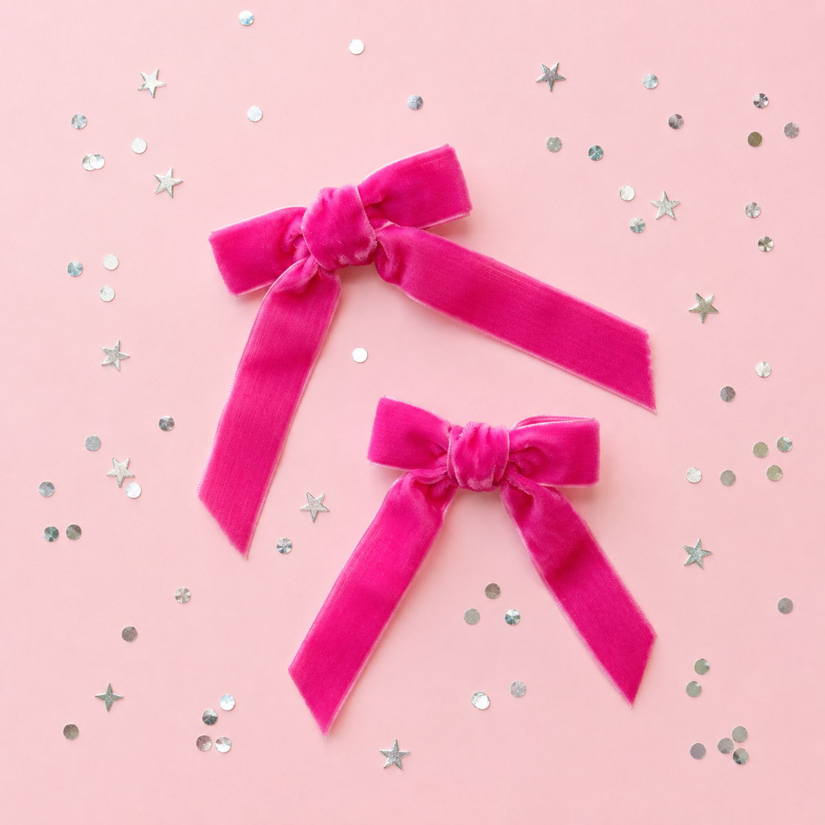 Two bright pink velvet hair bows arranged on a light pink background with scattered silver star and round confetti.
