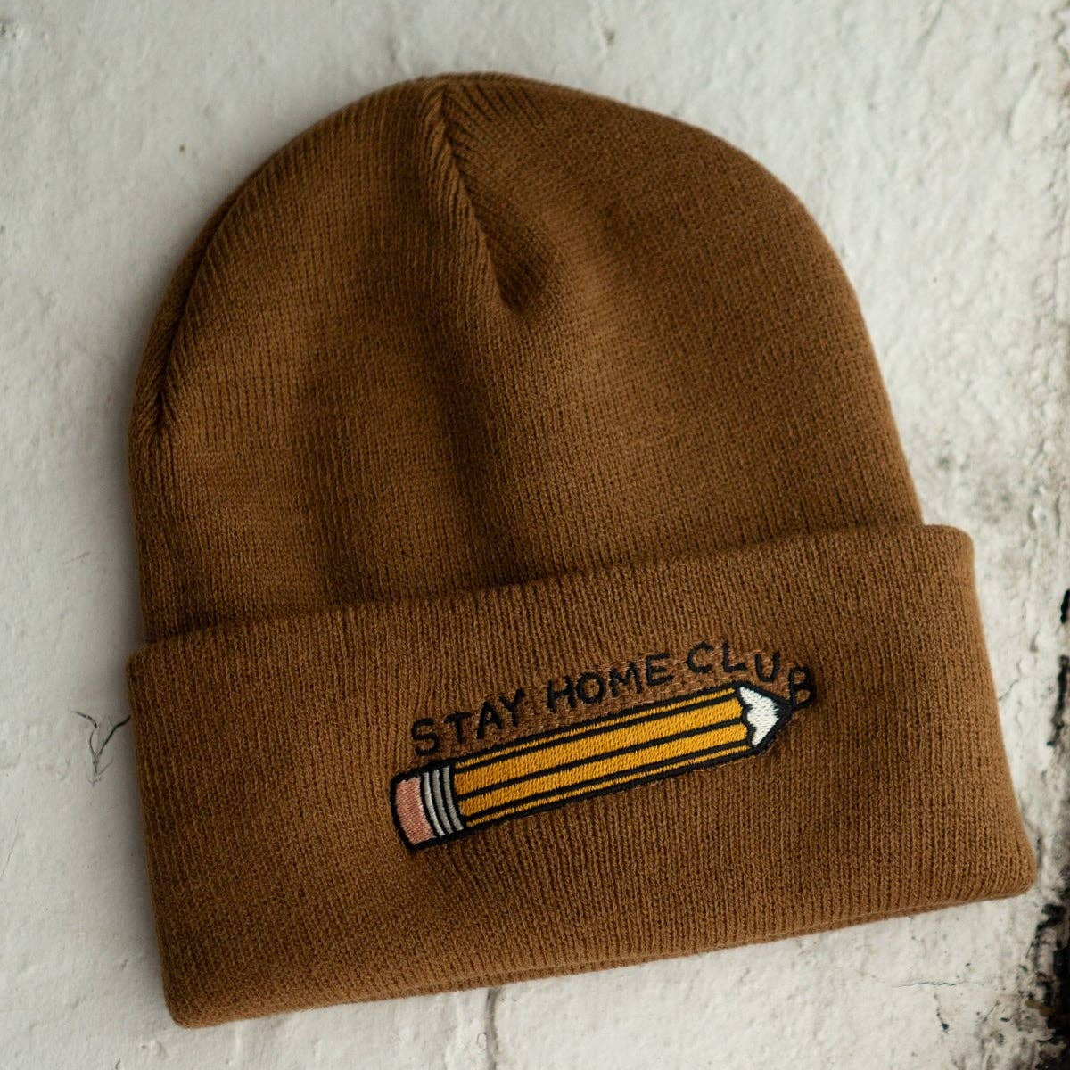 Brown beanie with a pencil and 'Stay Home Club' text on a textured white background