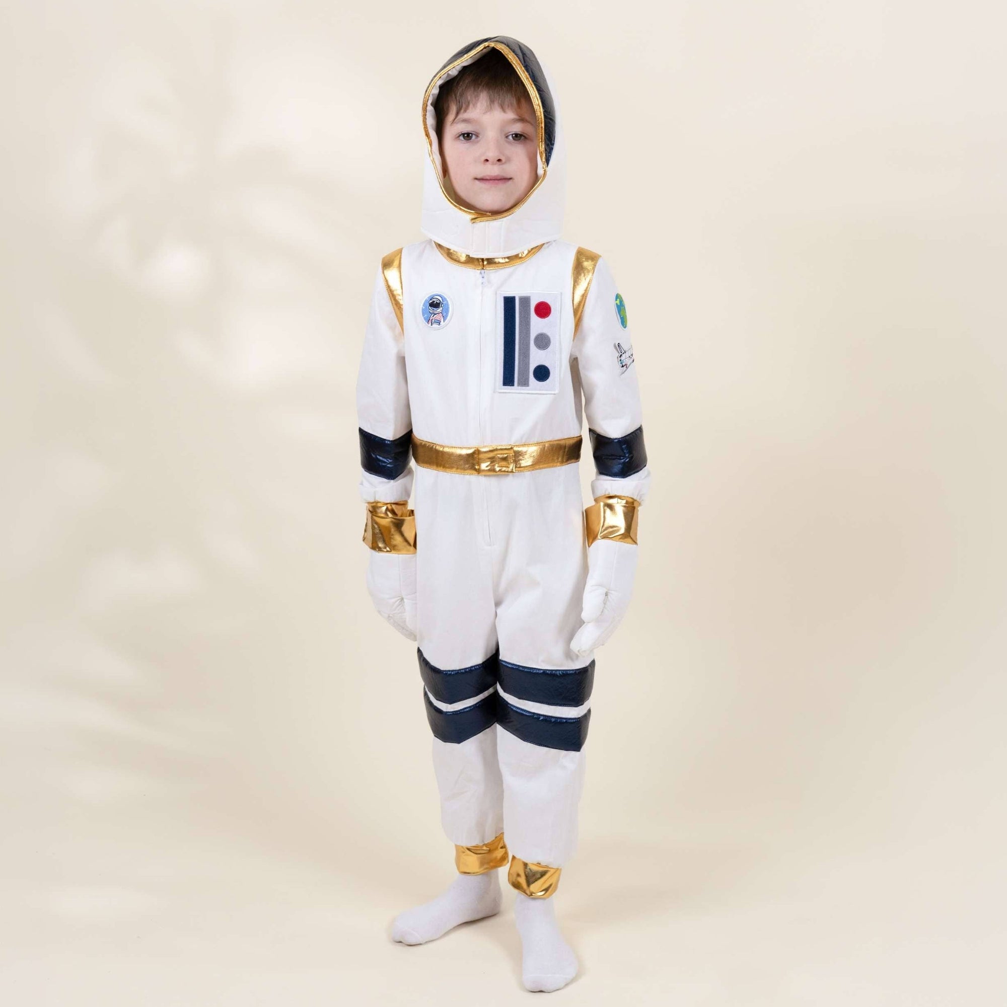 A child wearing a full-body astronaut costume stands against a plain warm beige studio background. The costume is predominantly white with gold trim and dark navy blue stripe accents on the sleeves, legs, and boots.