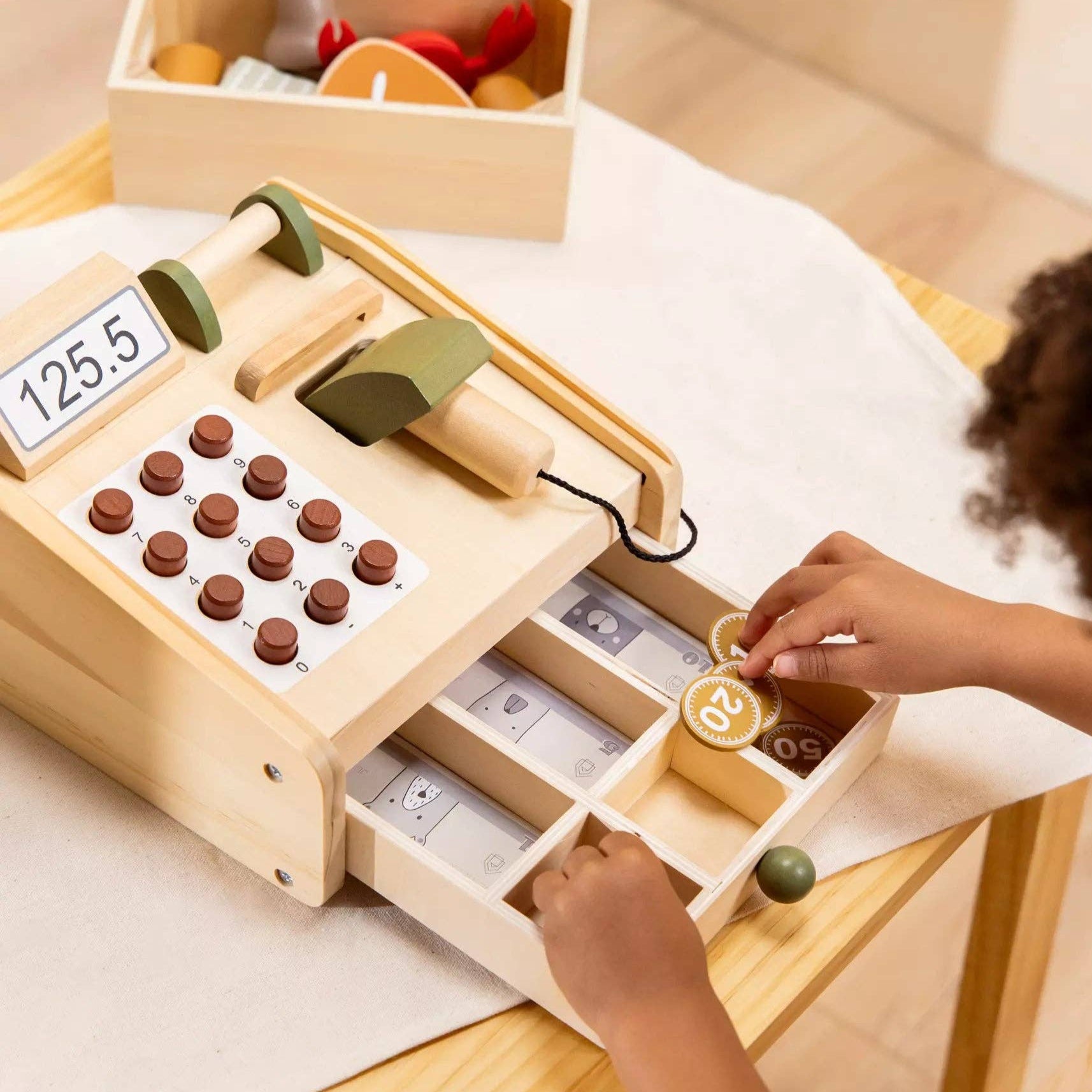 Child playing with a wooden toy cash register on a table.