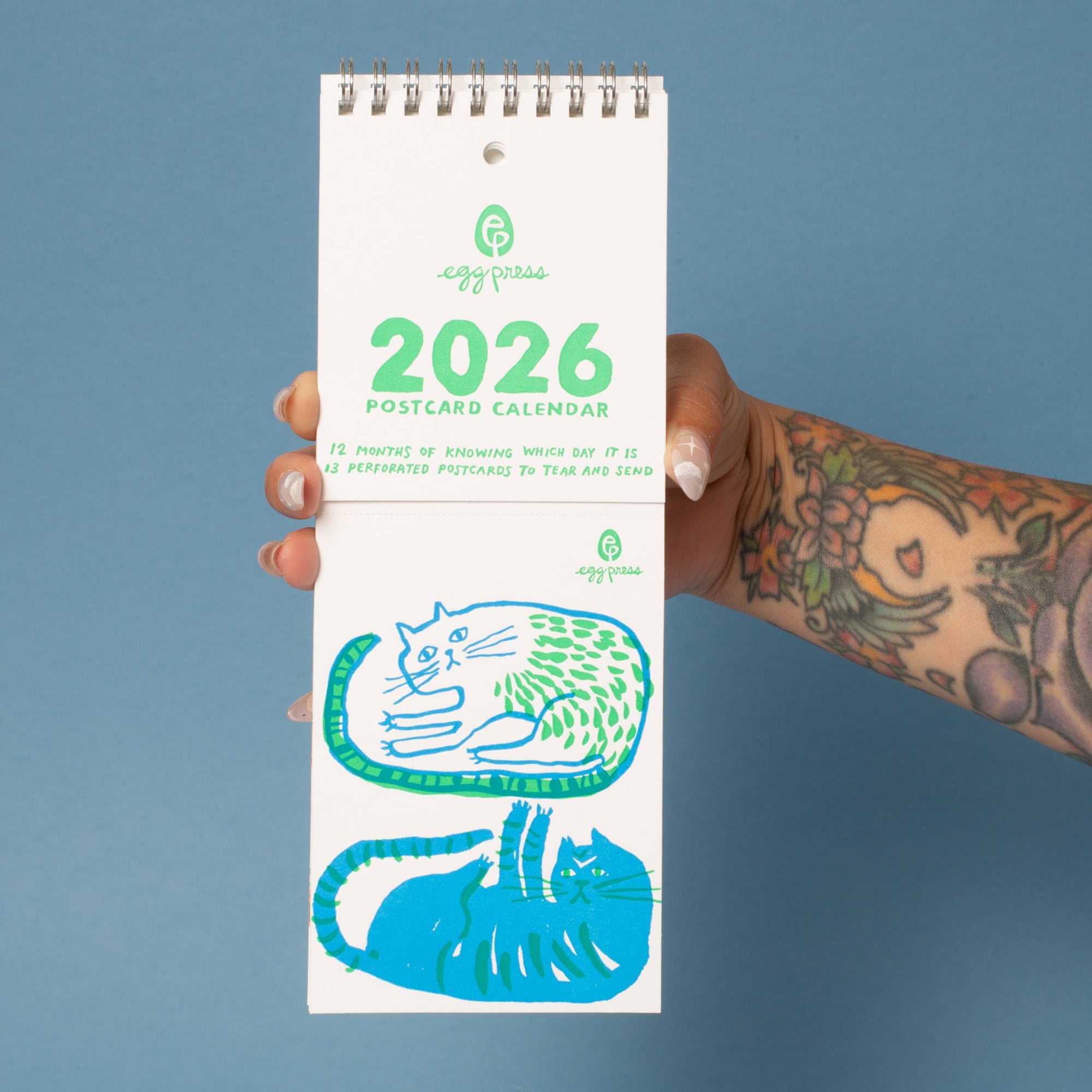 2026 postcard calendar held by a hand with tattoos on a blue background