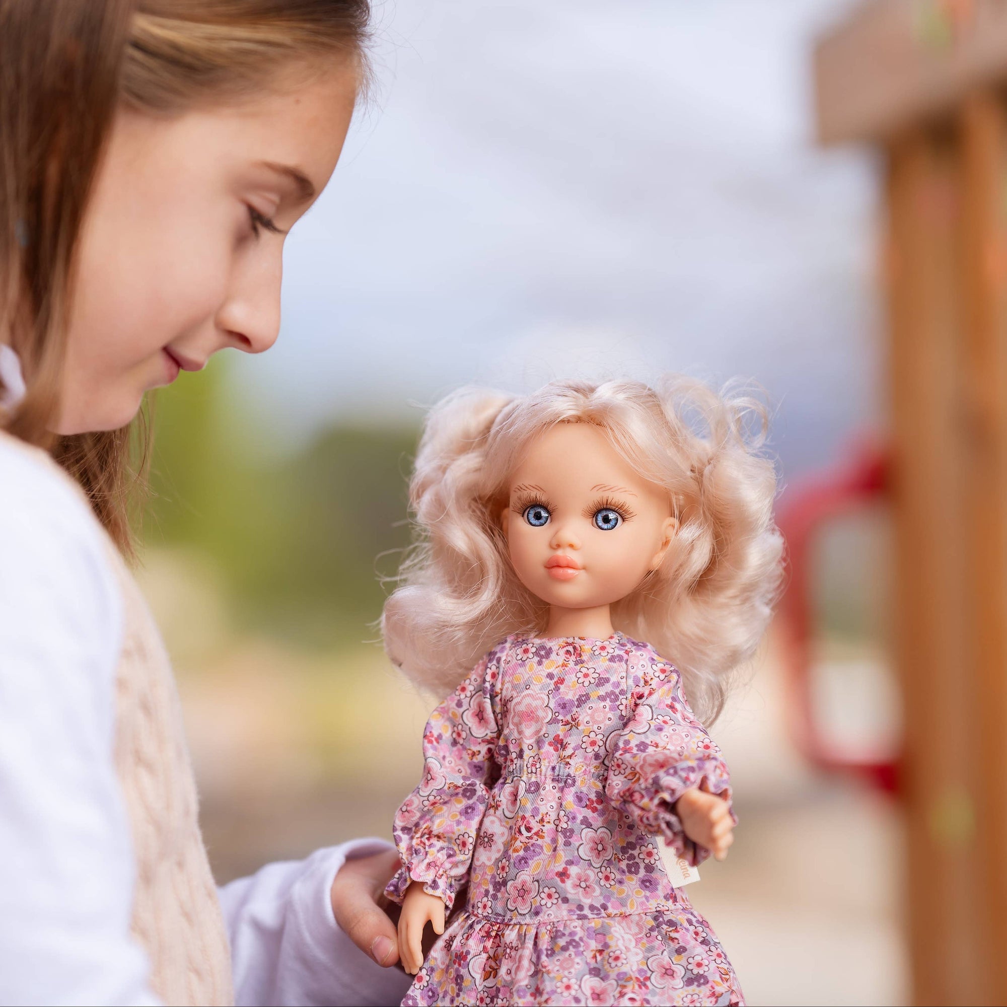 Young girl holding a doll with blonde hair and a floral dress outdoors.