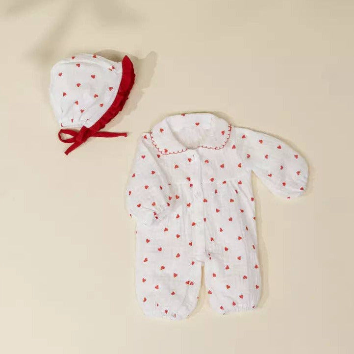 A baby footed one-piece sleeper in white muslin fabric with a red heart pattern, shown next to a matching bonnet with a red trim and bow, laid flat on a warm beige background.