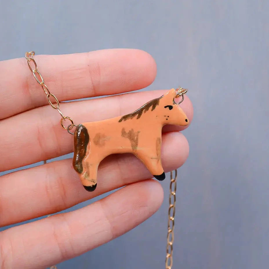 Hand holding a small horse-shaped pendant with a chain against a gray background