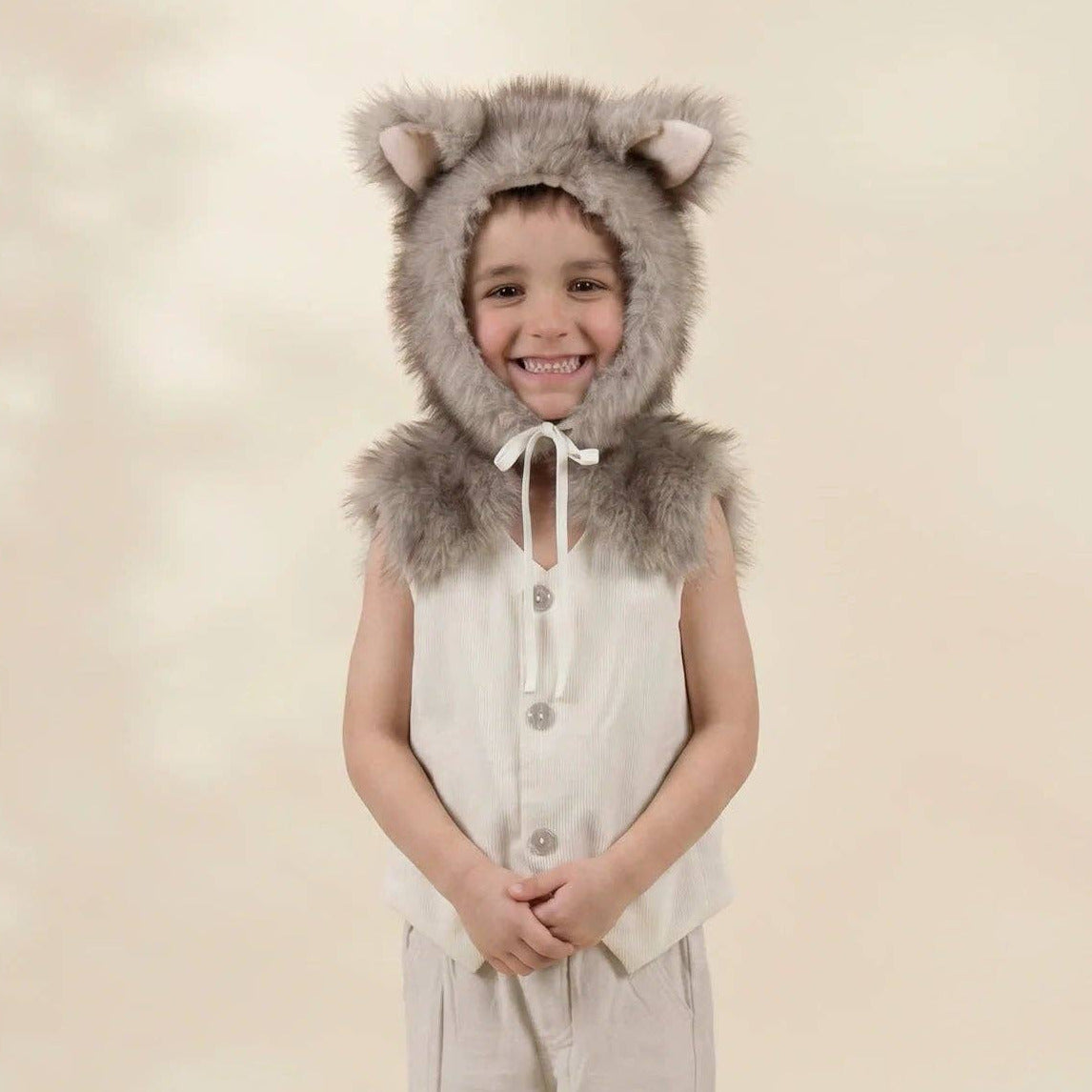 A young child modeling a wolf costume set featuring a gray faux-fur hood with ears, a sleeveless cream vest, matching beige pants, and gray slip-on shoes, set against a warm beige studio backdrop.