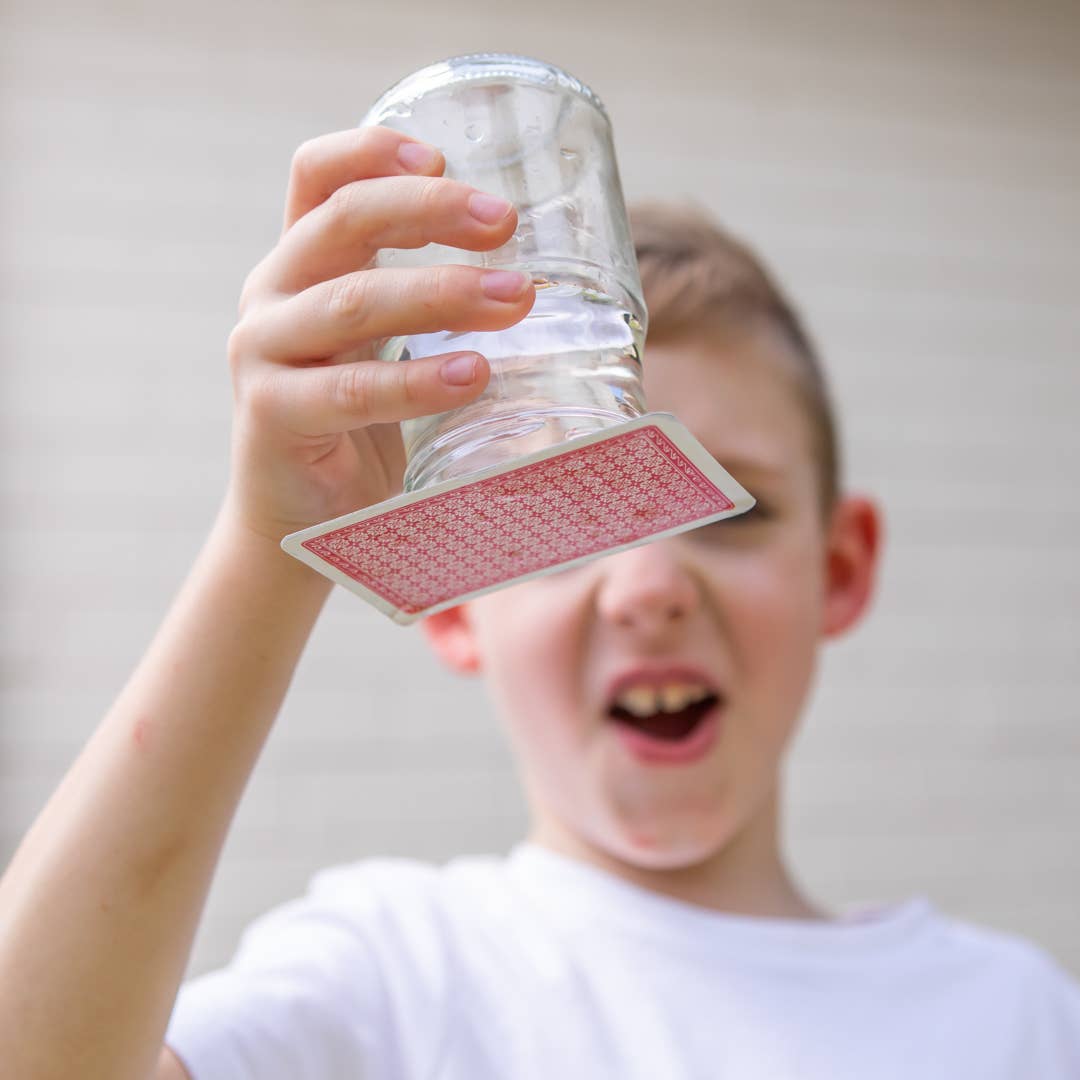 Child holding a clear plastic cup with a red playing card over their face against a plain background