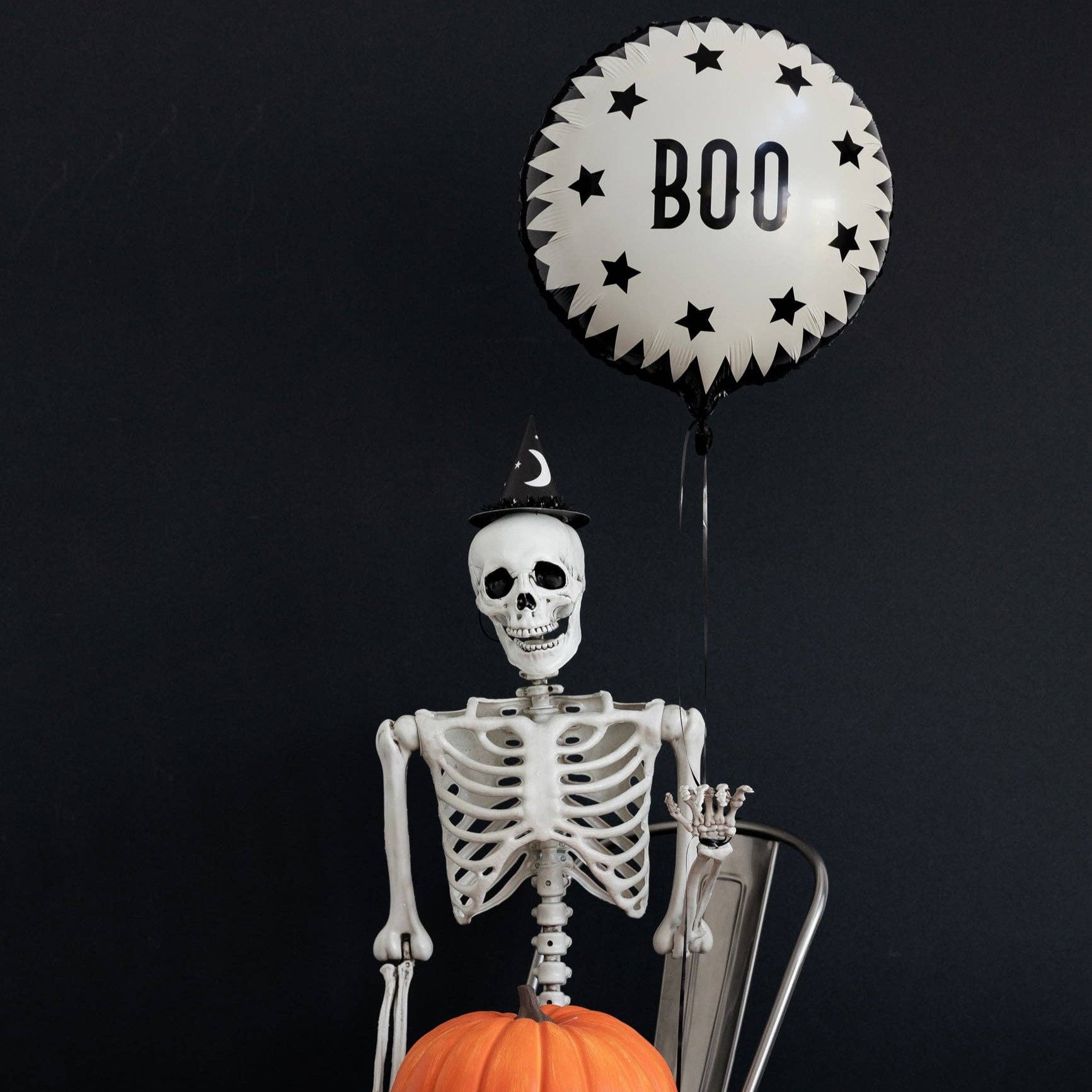 Skeleton holding a pumpkin with a 'BOO' balloon against a black background