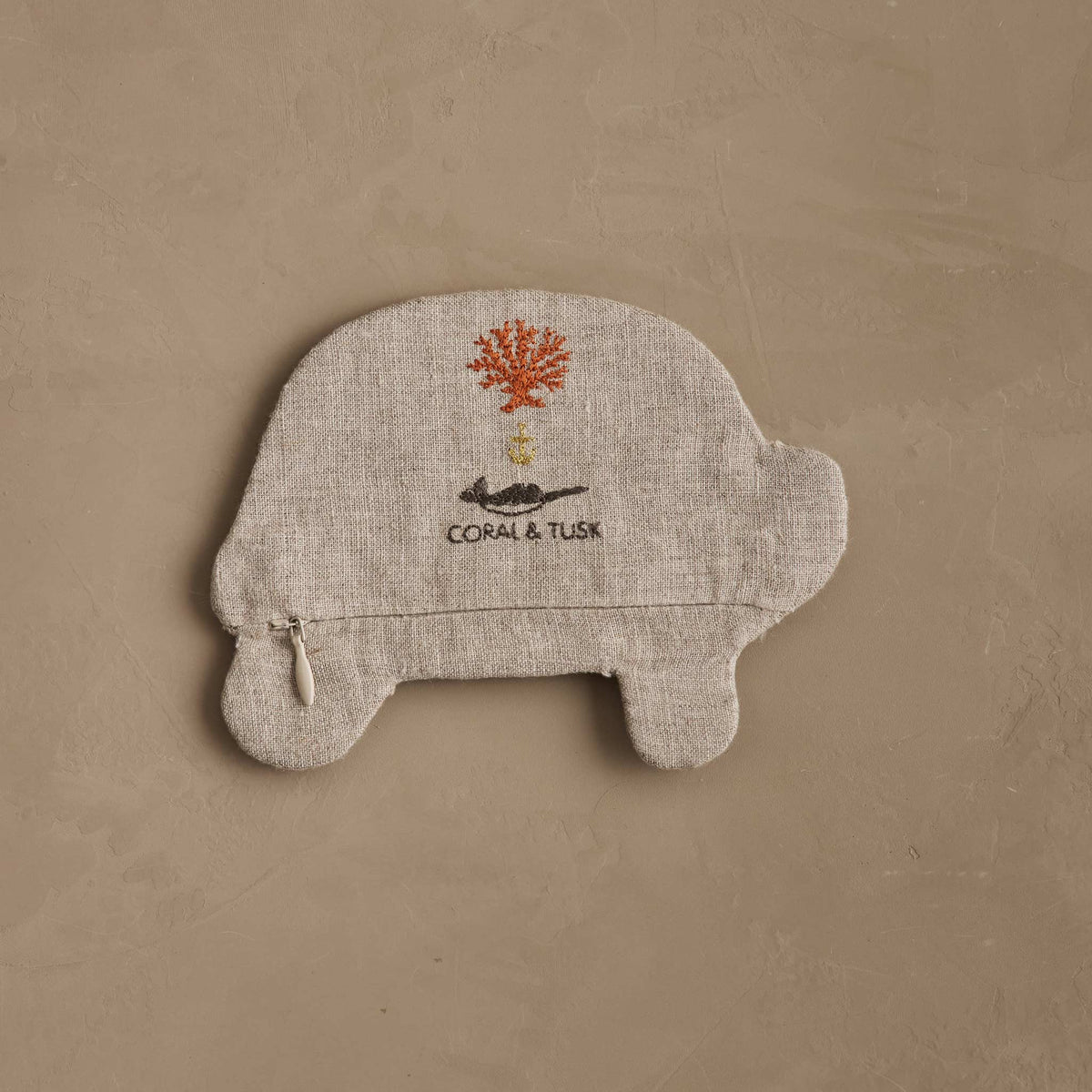 A plush car-shaped cushion in textured beige fabric, featuring an embroidered orange tree, a small black cat, and the text 'CORAL & TUSK' on the front, set against a matching beige textured background.