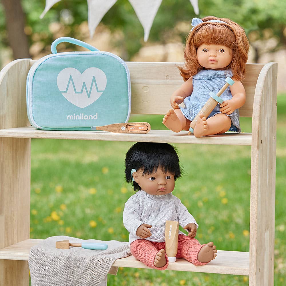Two dolls on a wooden shelf with a teal bag labeled 'miniland' in an outdoor setting.