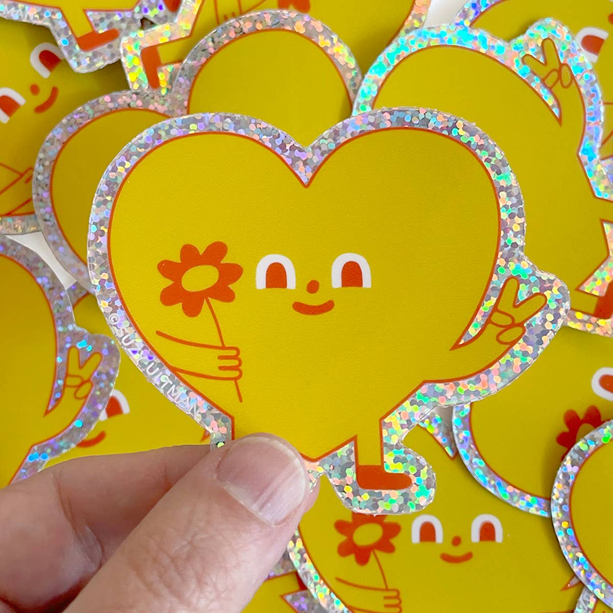 Hand holding a heart-shaped sticker with a face and flower, surrounded by more similar stickers on a yellow background.