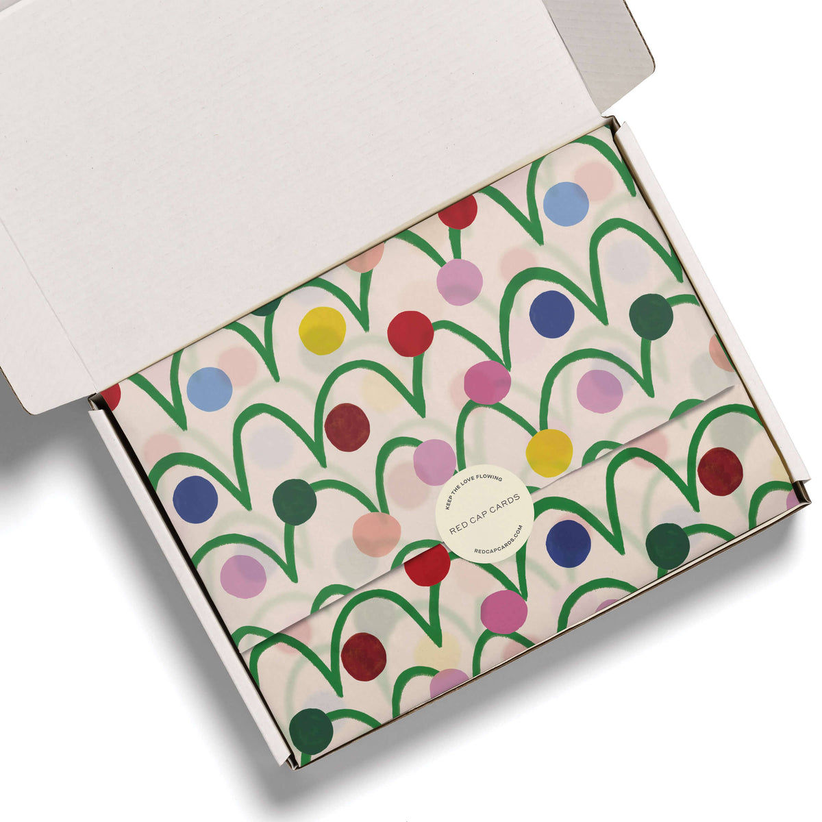 An open white cardboard gift box showing a folded envelope-style package wrapped in cream paper with a colorful abstract pattern of green lines and multicolored dots. A round white sticker on the front reads 'RED CAP Cards'.