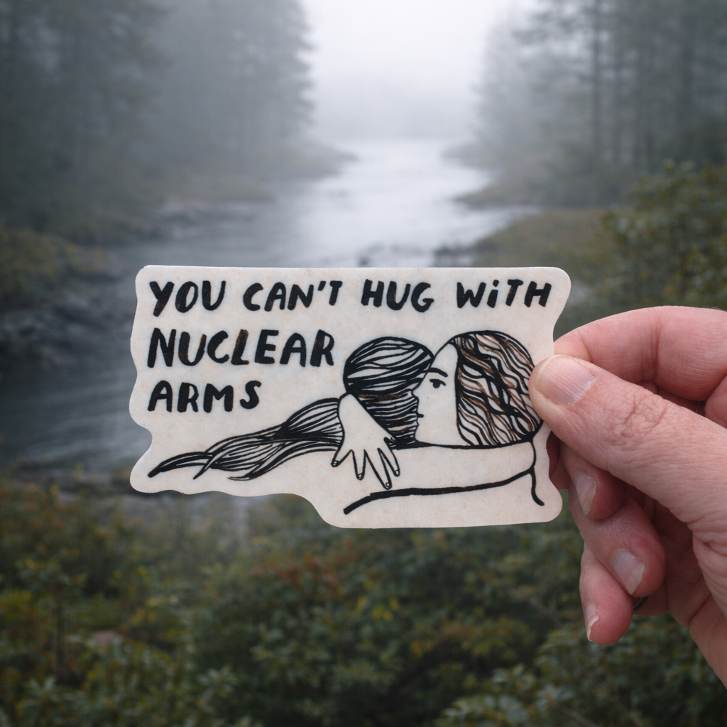 Hand holding a sticker with text 'You can't hug with nuclear arms' against a forest background